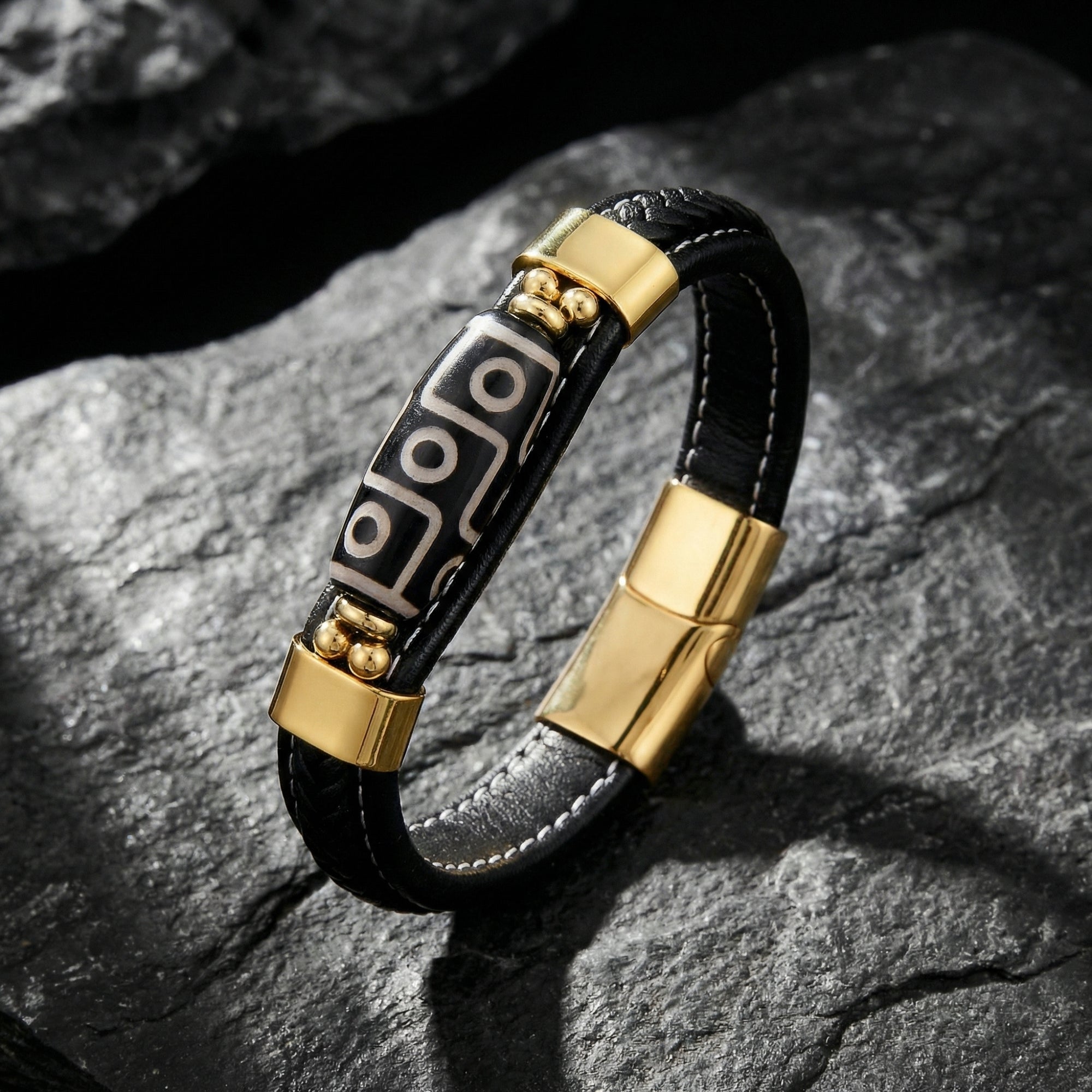 Men's black braided leather bracelet with gold PVD stainless steel hardware on a dark rocky background.
