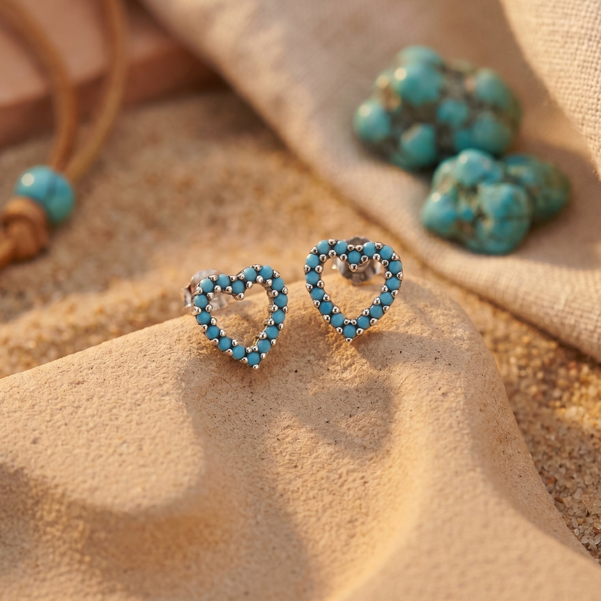 Silver heart-shaped turquoise stud earrings resting on a desert rock with raw turquoise stones.