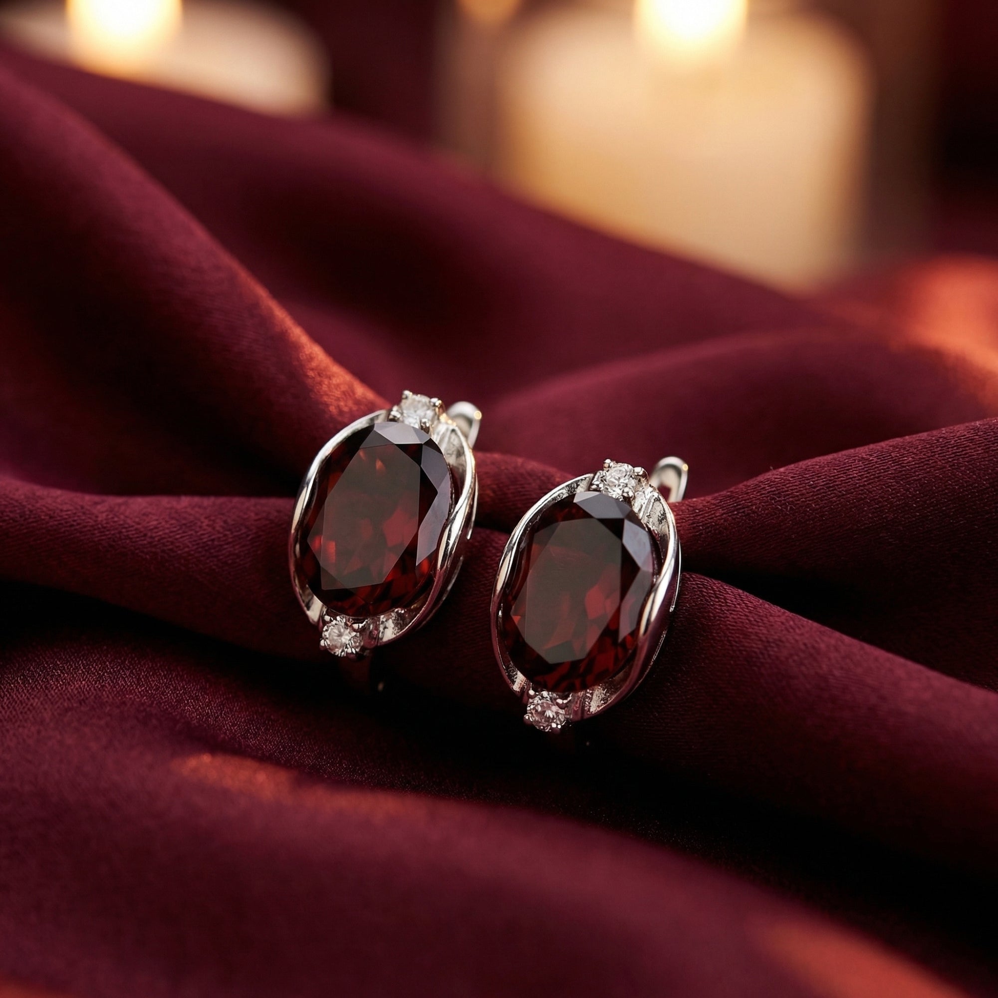 8x10mm oval cut garnet drop earrings in sterling silver resting on burgundy silk with a warm candlelit background.