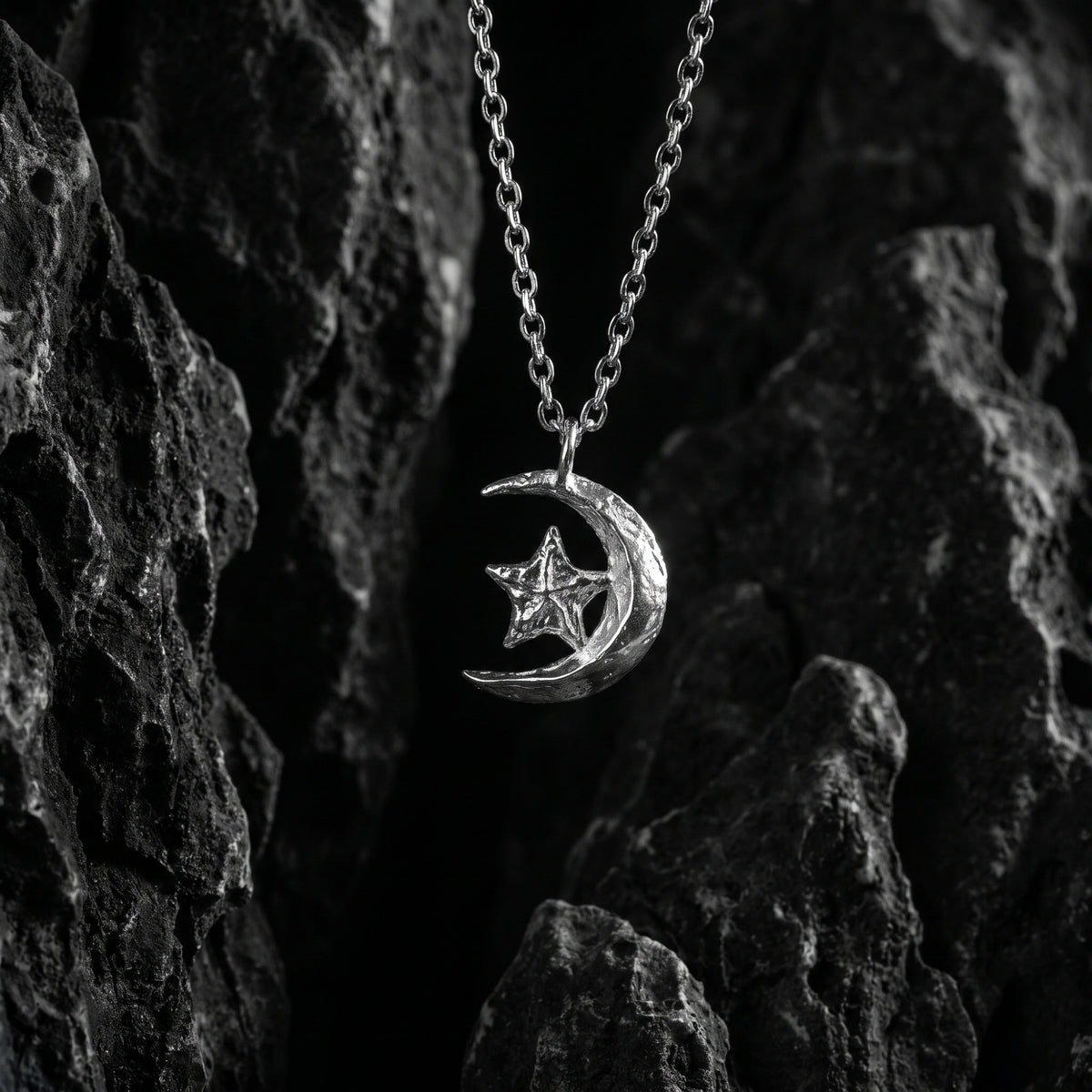 Silver crescent moon and star pendant hanging on a chain among dark textured rocks.