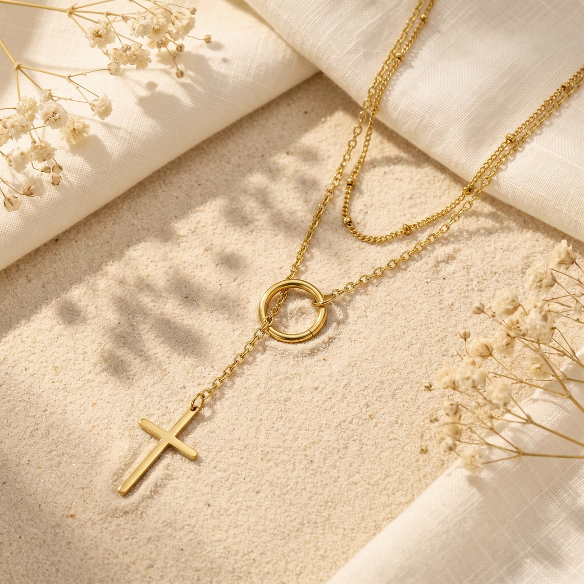 Gold-tone layered cross lariat necklace displayed flat on fine sand with cream linen and dried flowers.