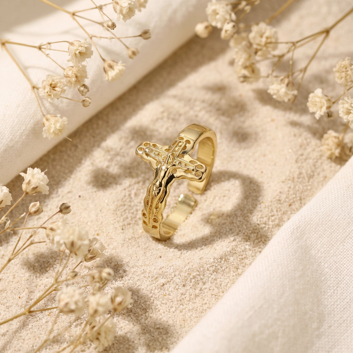 Gold-tone sculpted cross ring standing upright on light sand among white dried flowers.