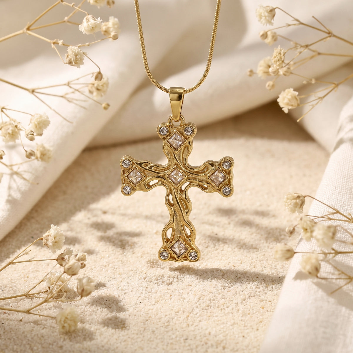 Gold-tone ornate filigree cross necklace with clear stone accents, suspended over sand among white flowers and fabric.