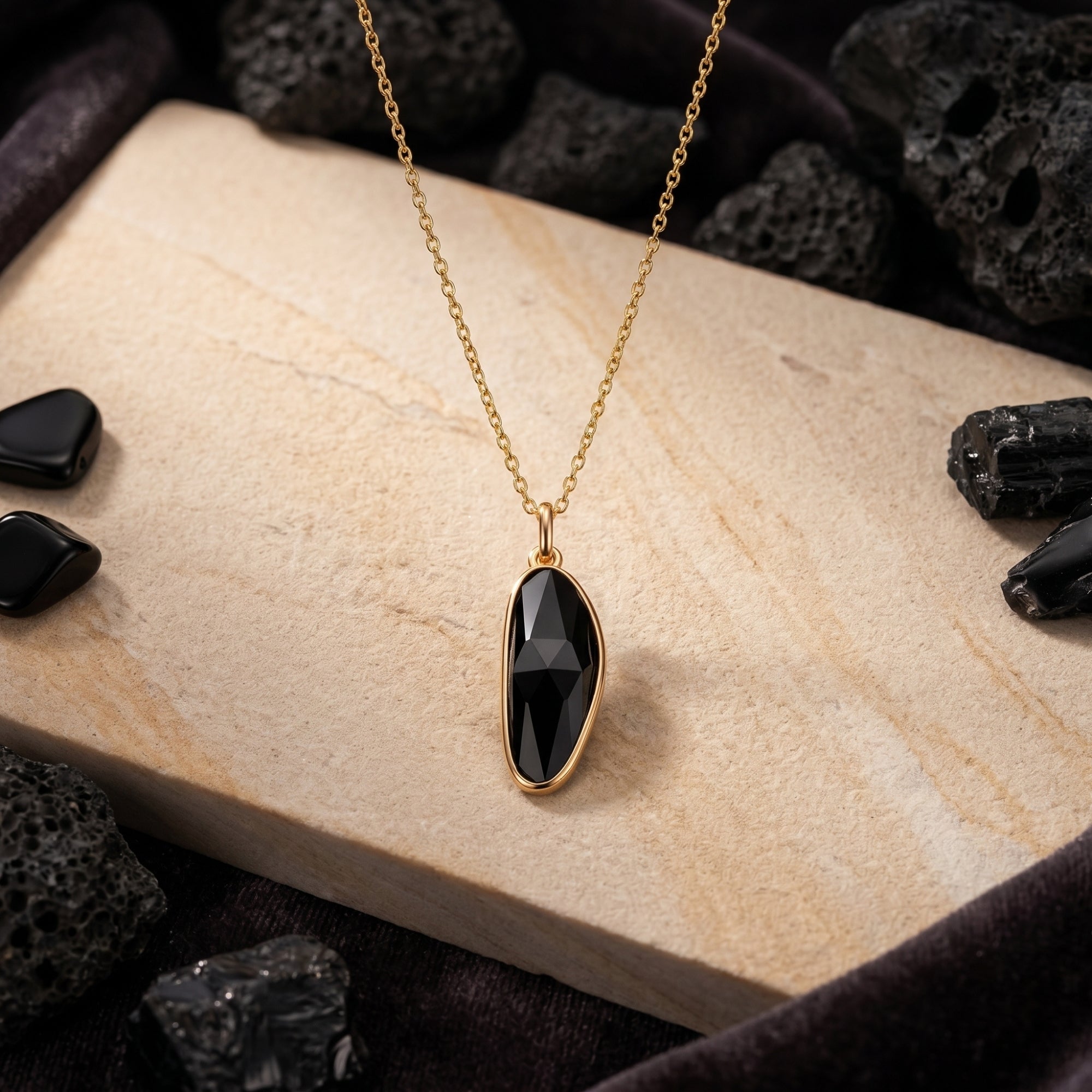 Faceted irregular-cut black onyx pendant on a gold chain, styled on a stone slab with volcanic rocks.