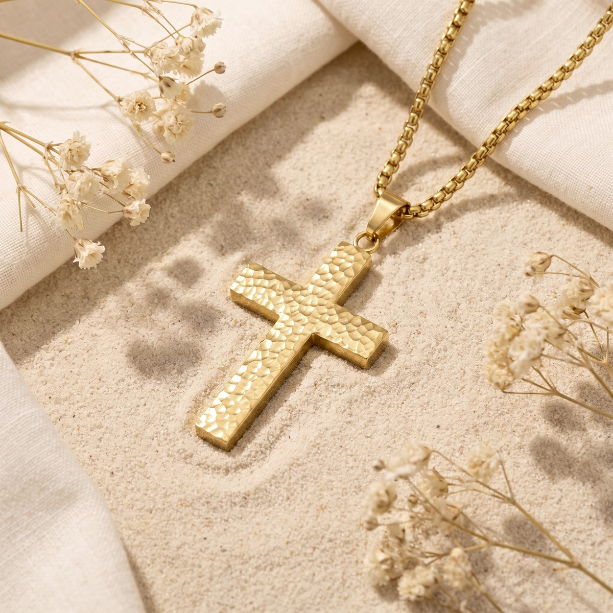 Gold-tone hammered cross pendant with box chain resting on sand beside dried flowers and cream fabric.