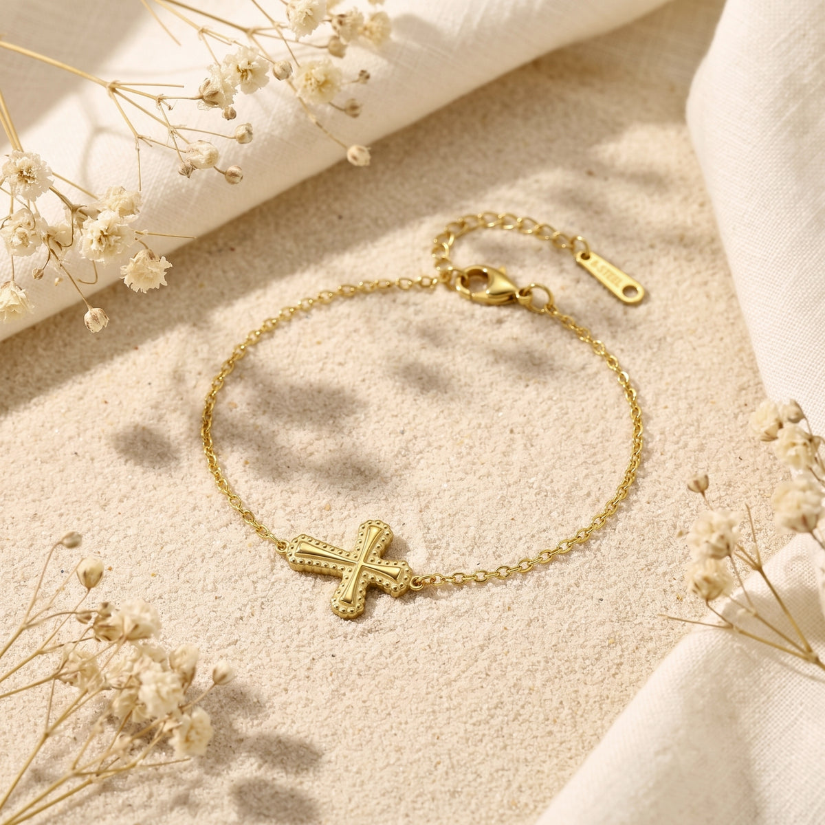 Gold-toned beaded cross bracelet displayed flat on textured sand with dried white flowers.
