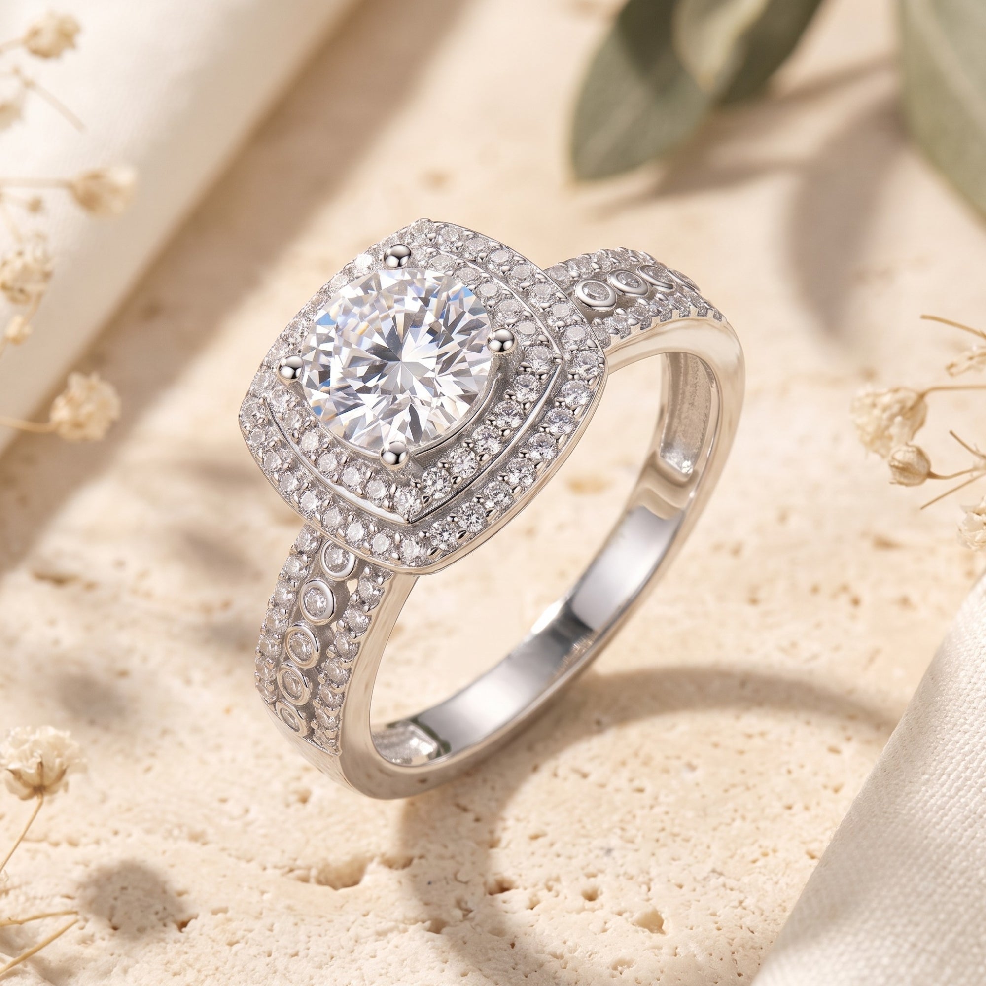 Silver cushion double halo ring with round brilliant stone on a textured stone surface with dried flowers.