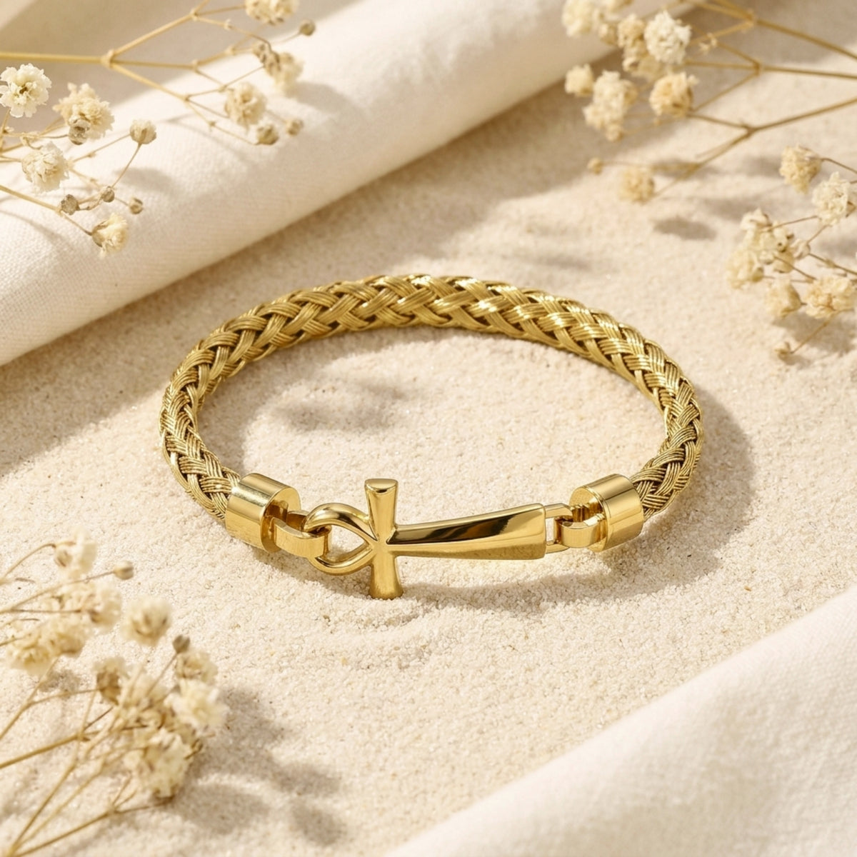 Gold-tone braided bracelet lying flat on sand with its Ankh clasp facing forward.