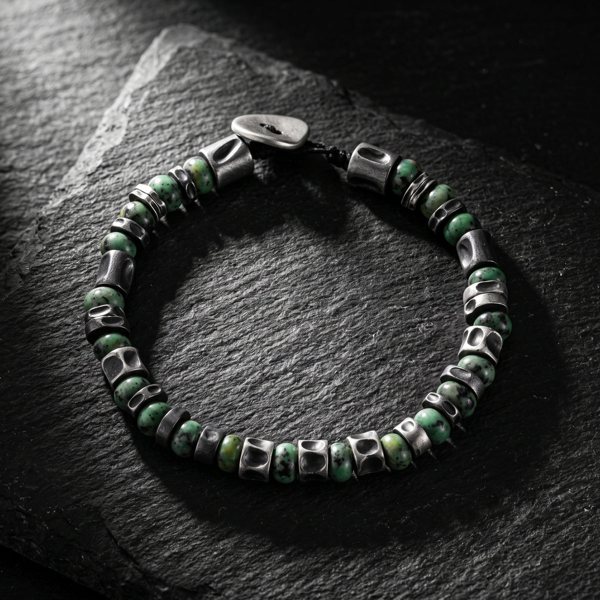Full lay-flat view of the green stone bead and silver barrel spacer bracelet on dark slate.