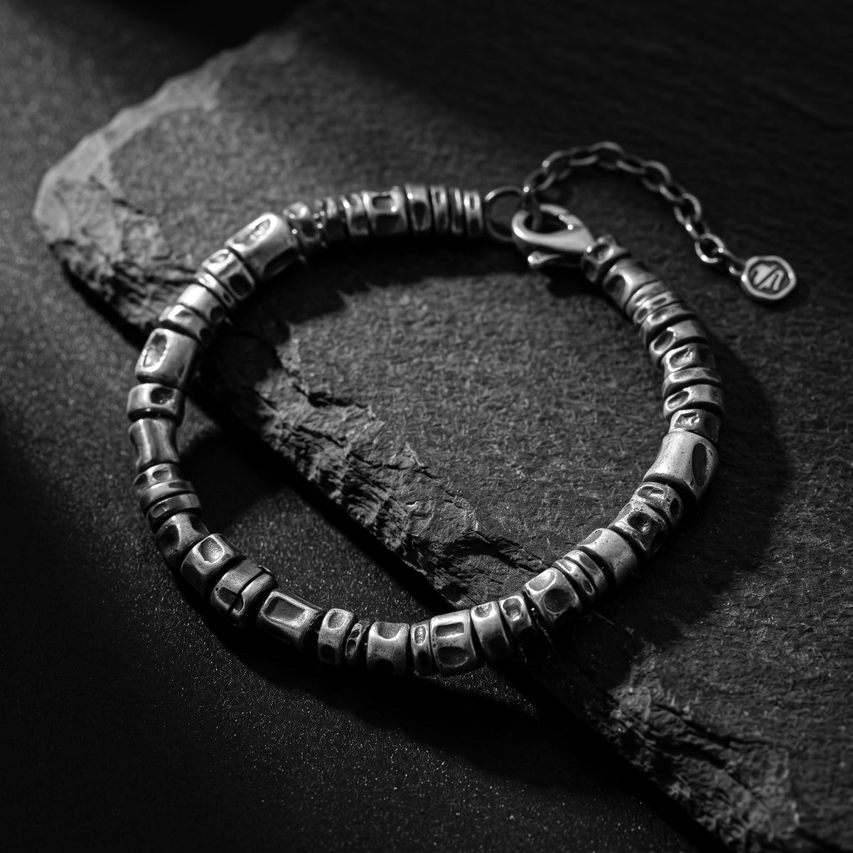 Textured silver barrel-link bracelet with an oxidized finish arranged on a slate slab over a dark surface.
