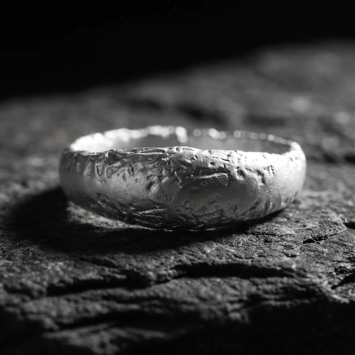 Close-up of a textured silver open-band ring resting horizontally on a dark textured surface.