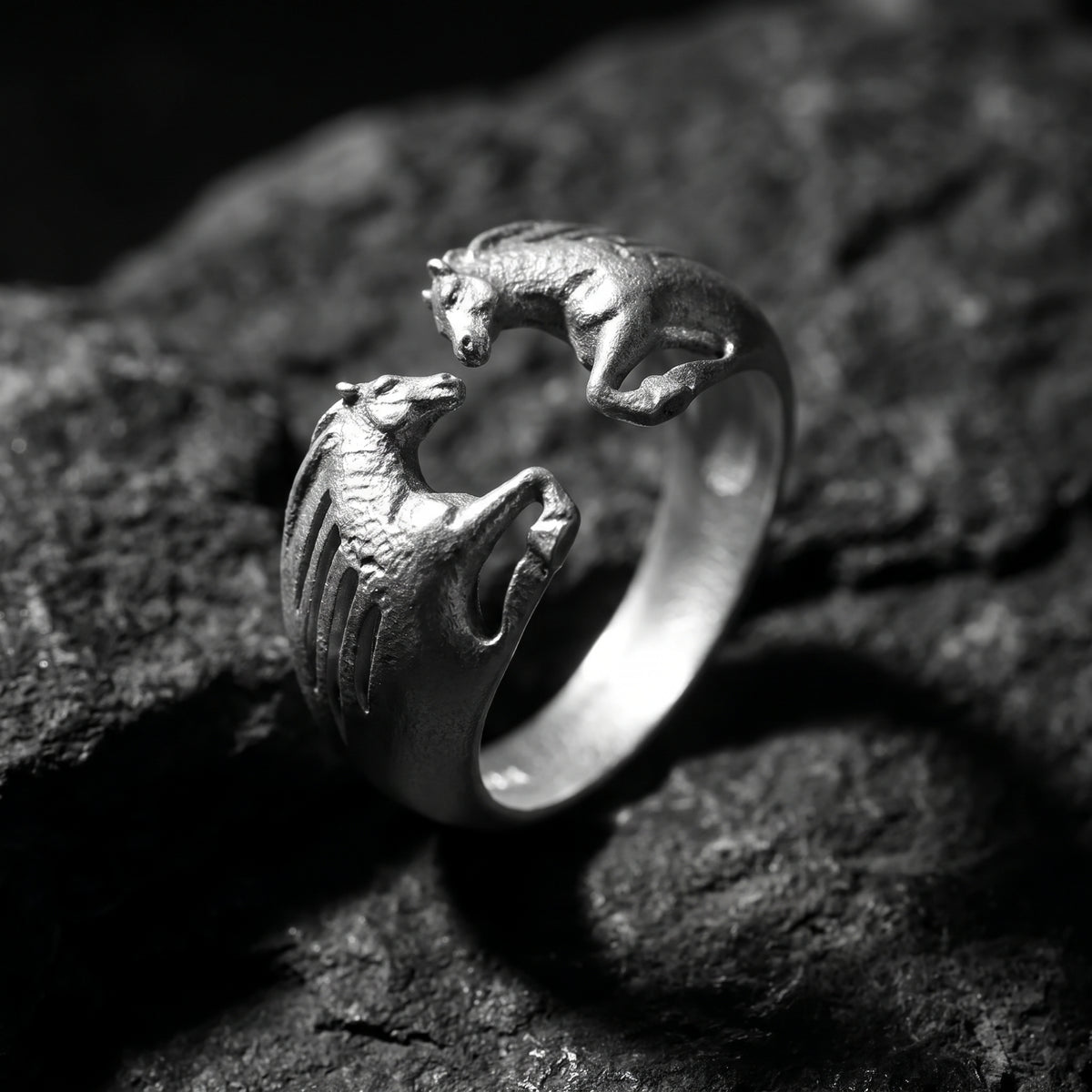 Close-up of the silver dual horse open ring resting on dark rock, with two sculpted horses facing each other.