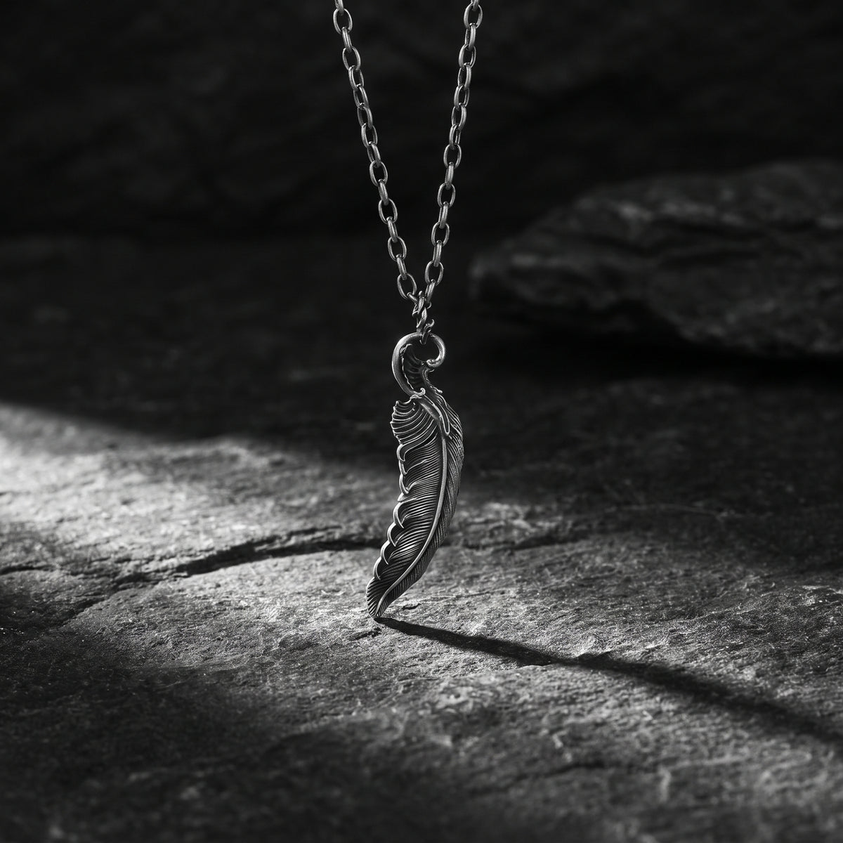 Silver feather pendant hanging from its chain with the tip touching a dark textured stone surface.