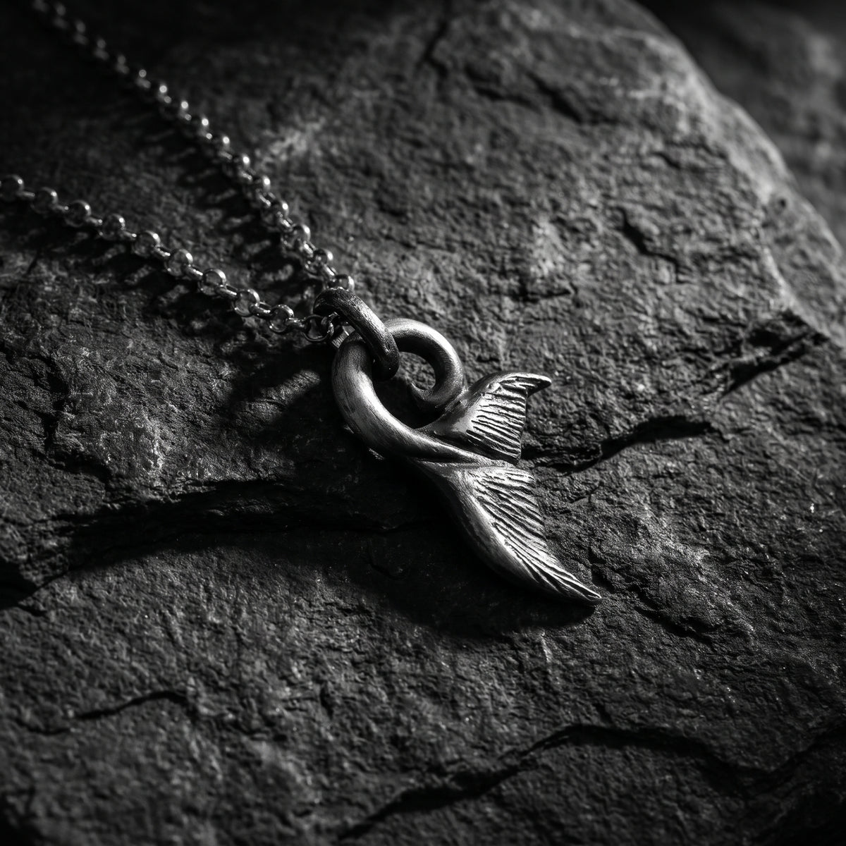 Close-up of a textured silver whale tail fluke pendant and chain lying on a dark slate surface.