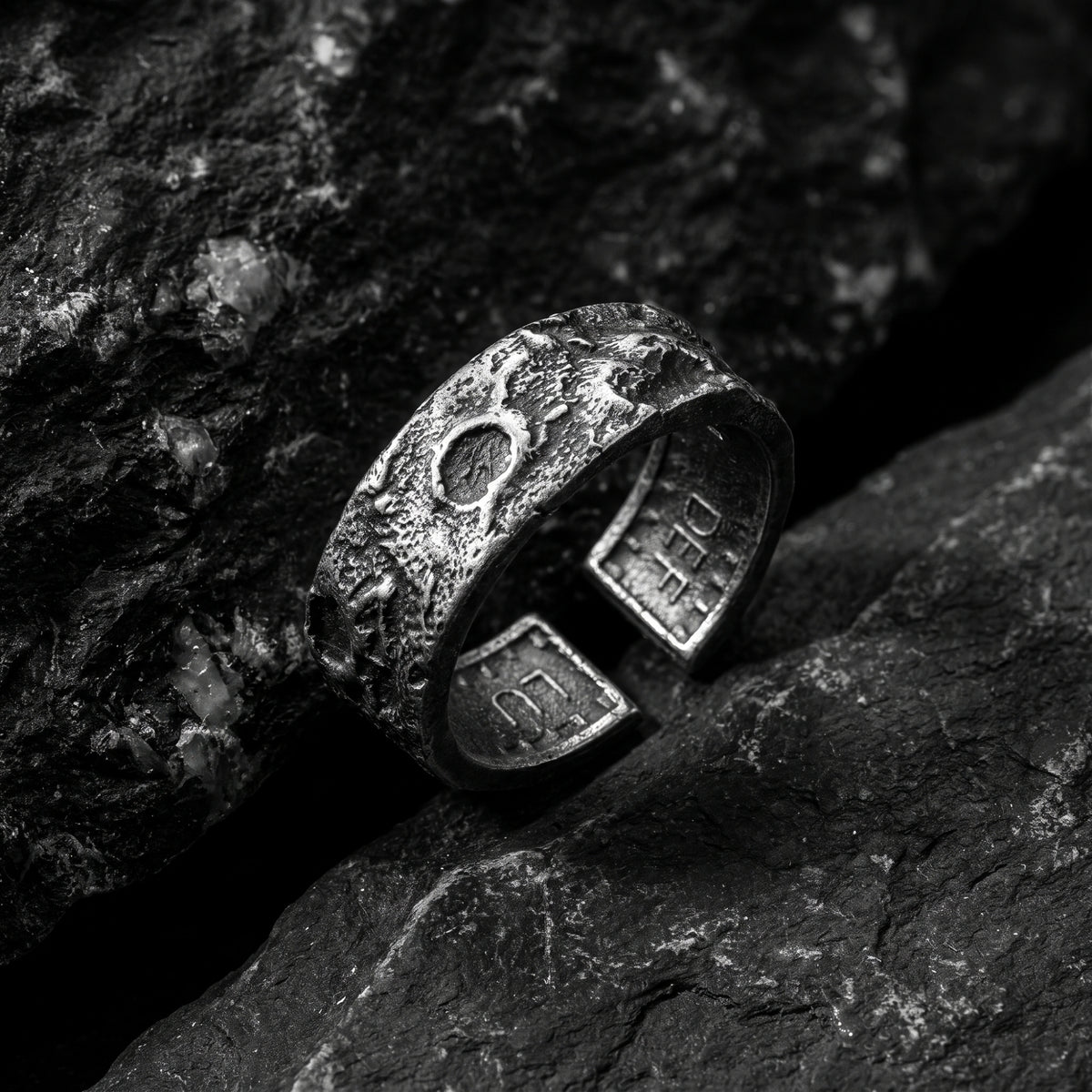 Textured silver open-band ring resting on dark textured rocks in an angled close-up.