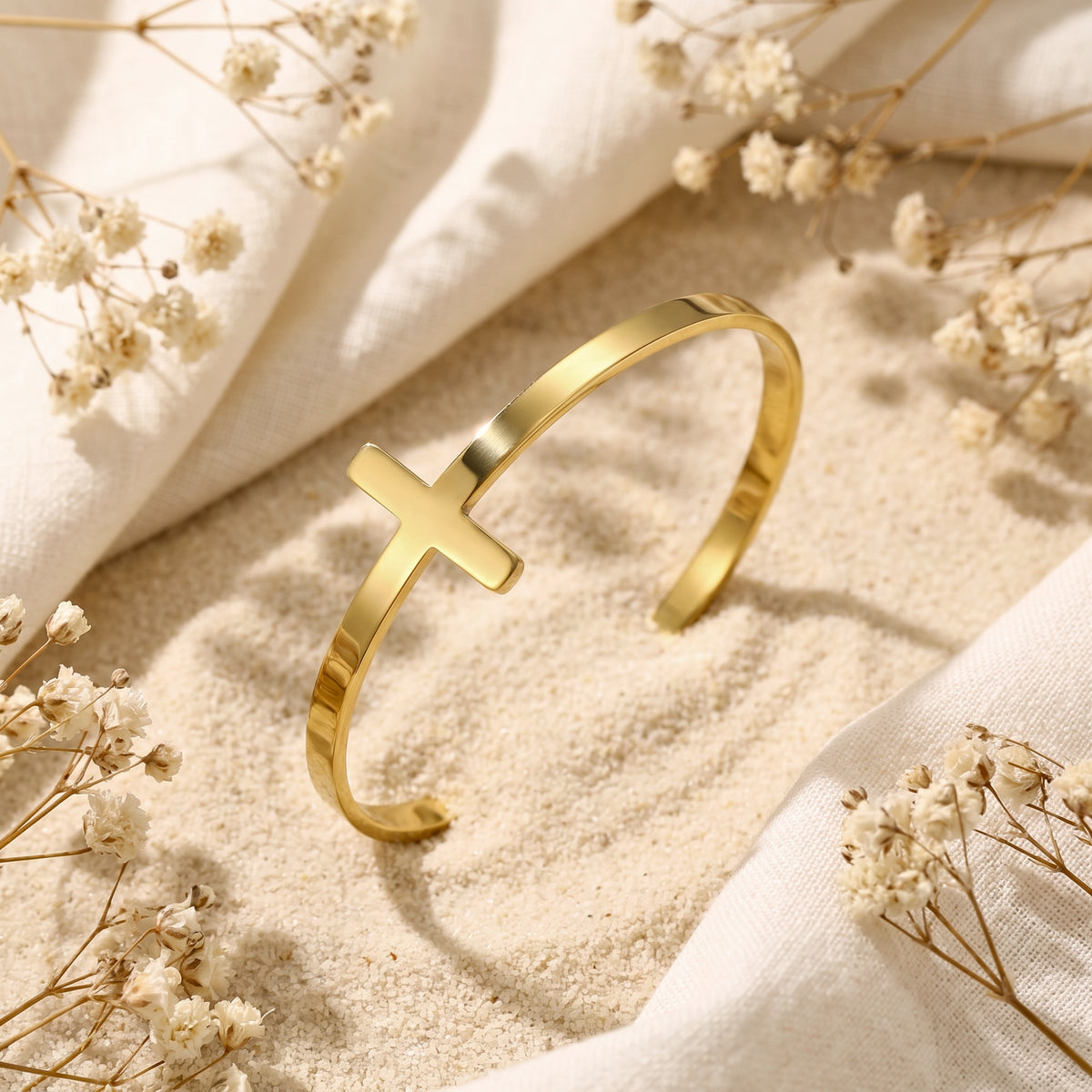 Gold-tone cross cuff bracelet resting at an angle in pale sand beside dried flowers and cream fabric.