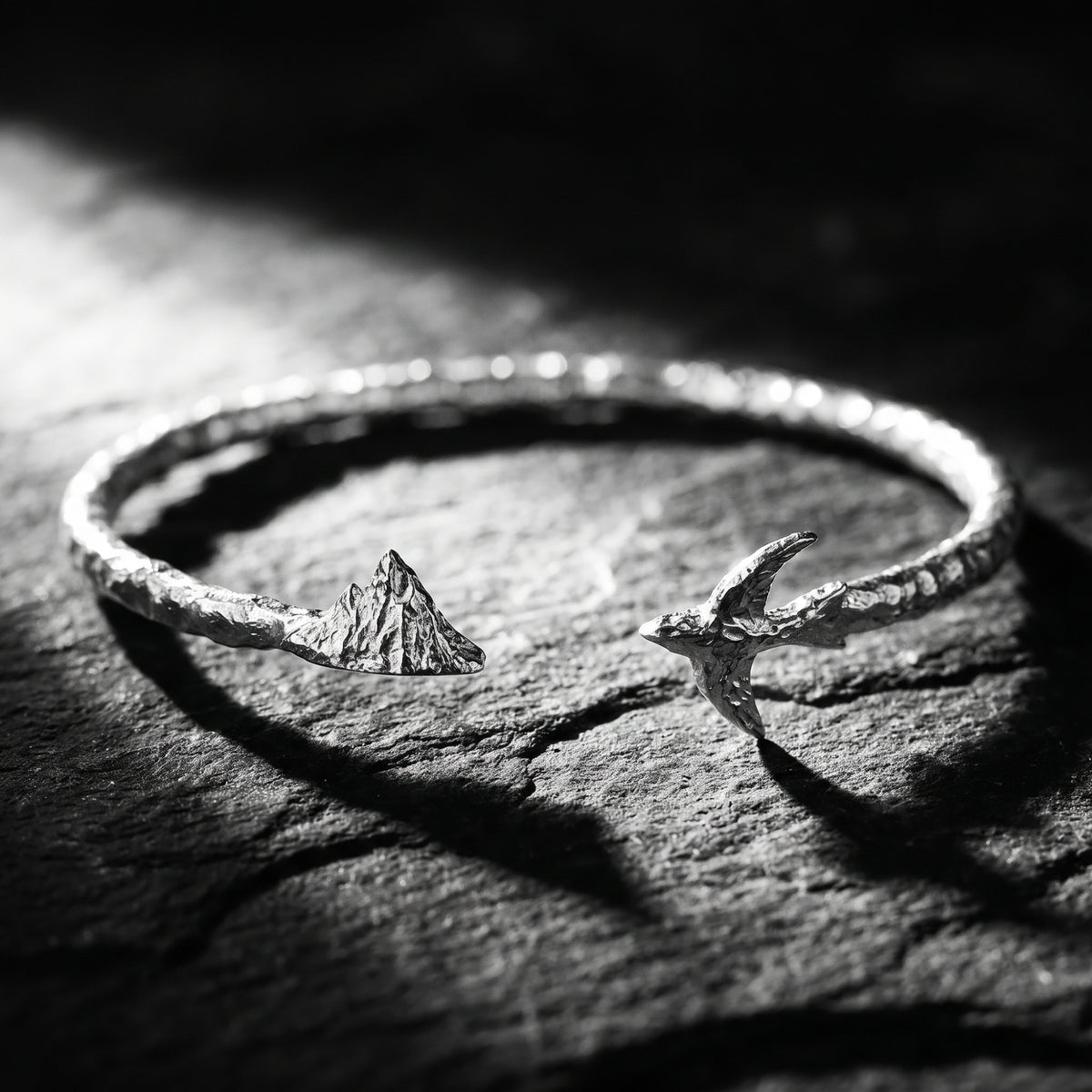Textured silver open cuff bracelet on dark rock, with sculpted mountain and bird ends facing across the gap.