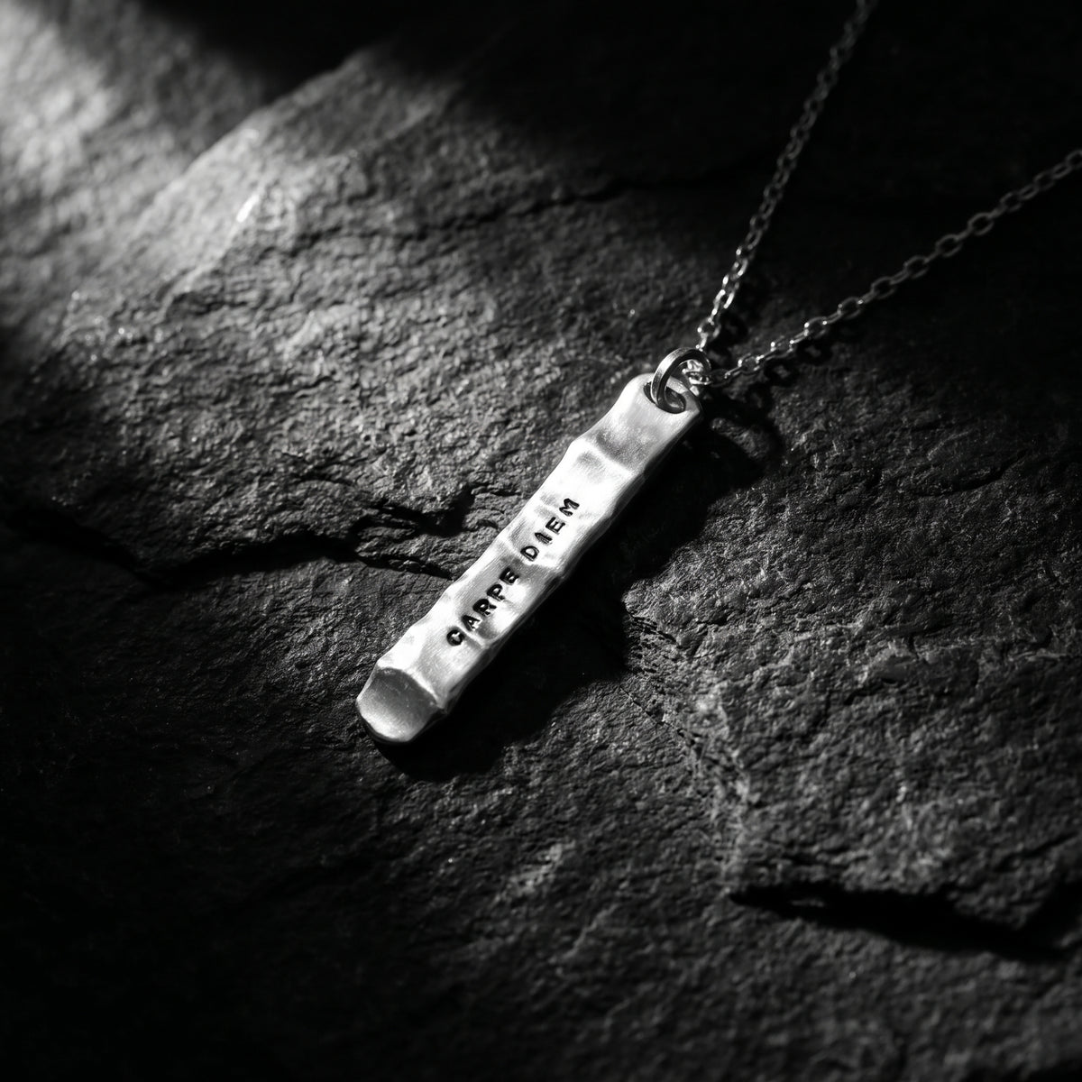 Silver bar pendant stamped with CARPE DIEM, lying at an angle on dark slate.