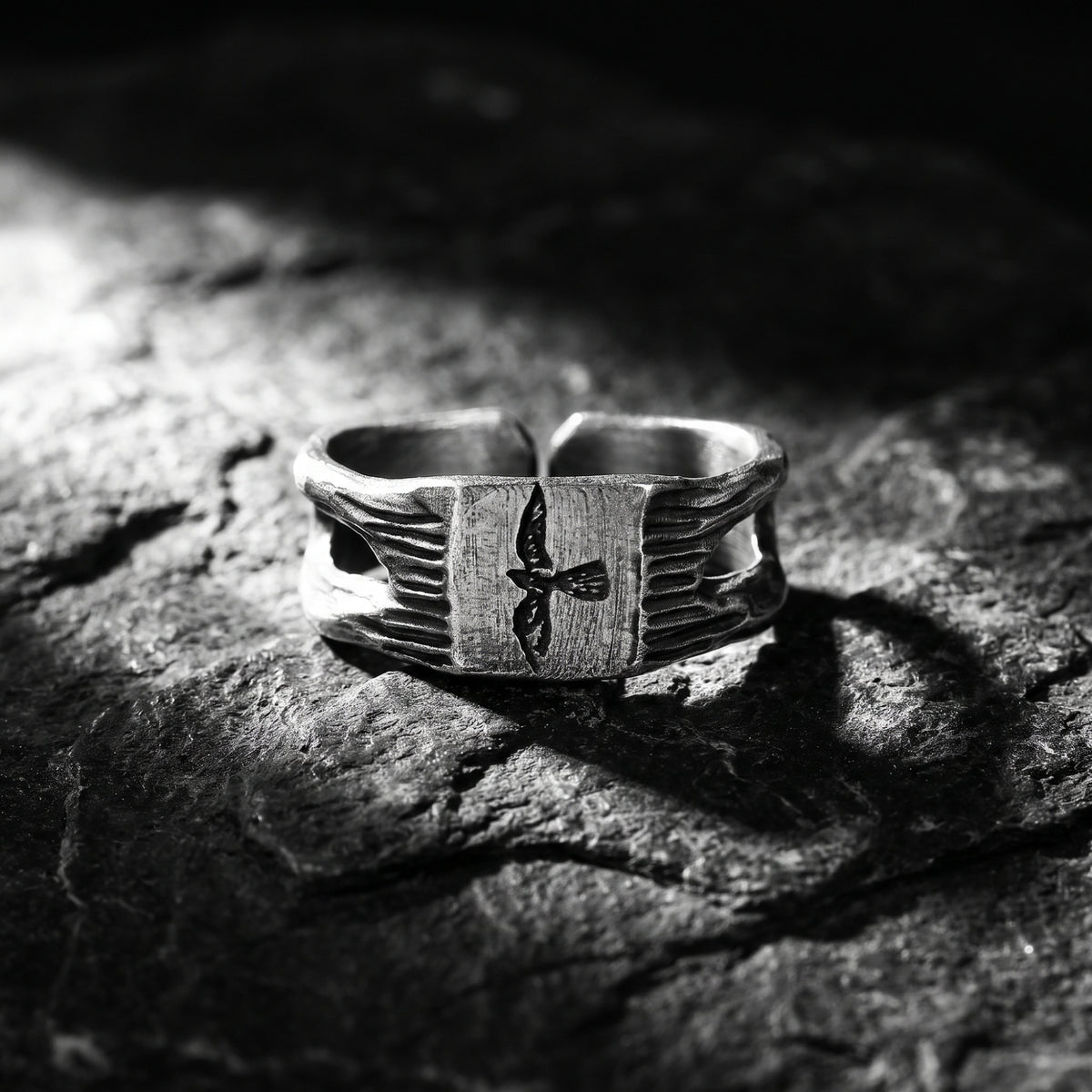 Silver signet ring with a carved falcon motif resting on dark textured stone, front view.