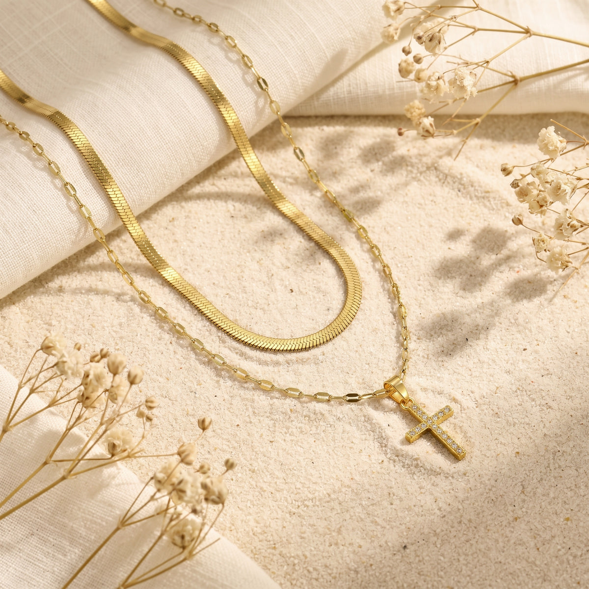 Flat lay of the gold-tone layered cross necklace with herringbone chain and pendant on a sand and linen surface.