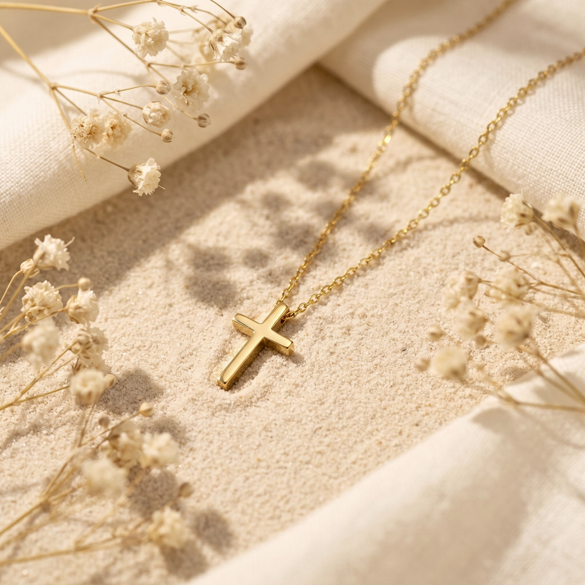 Gold-tone cross pendant necklace lying on sand among cream fabric and dried white flowers.