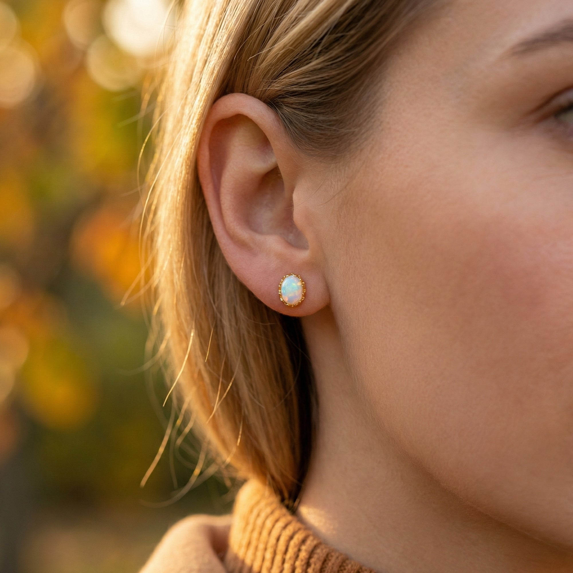 A woman wearing a yellow gold Novalie stud earring with an oval white opal in an outdoor setting.