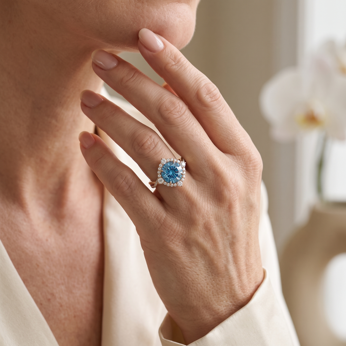Round blue moissanite halo ring in rose gold worn on a model's hand touching her chin.