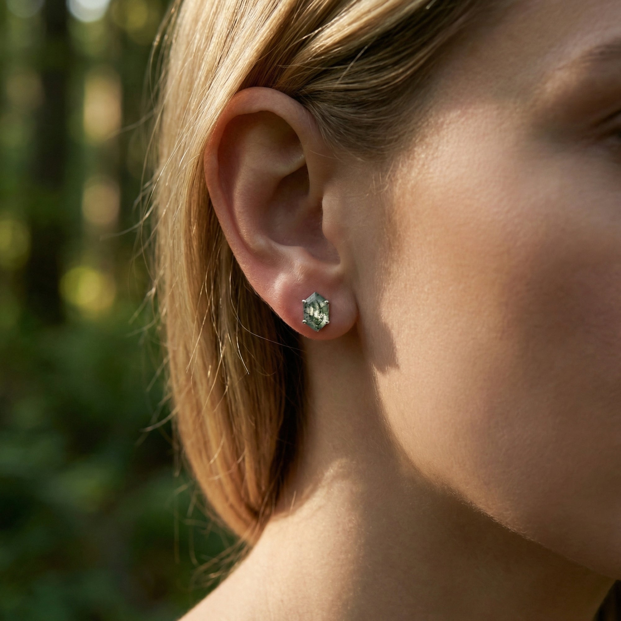 Sterling silver hexagon moss agate stud earring worn on a woman's ear with a blurred forest background.