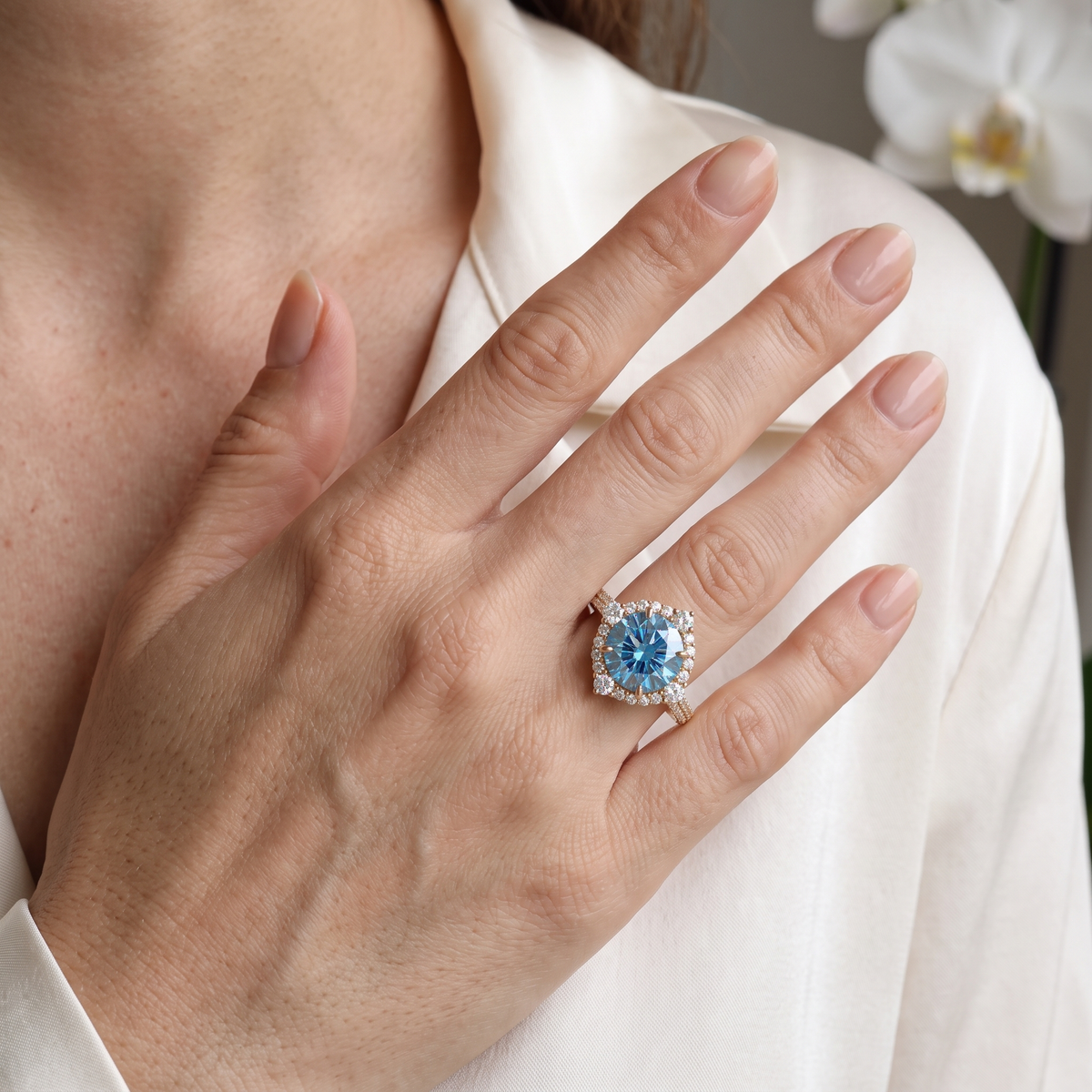 Round blue moissanite halo engagement ring in rose gold worn on a hand.