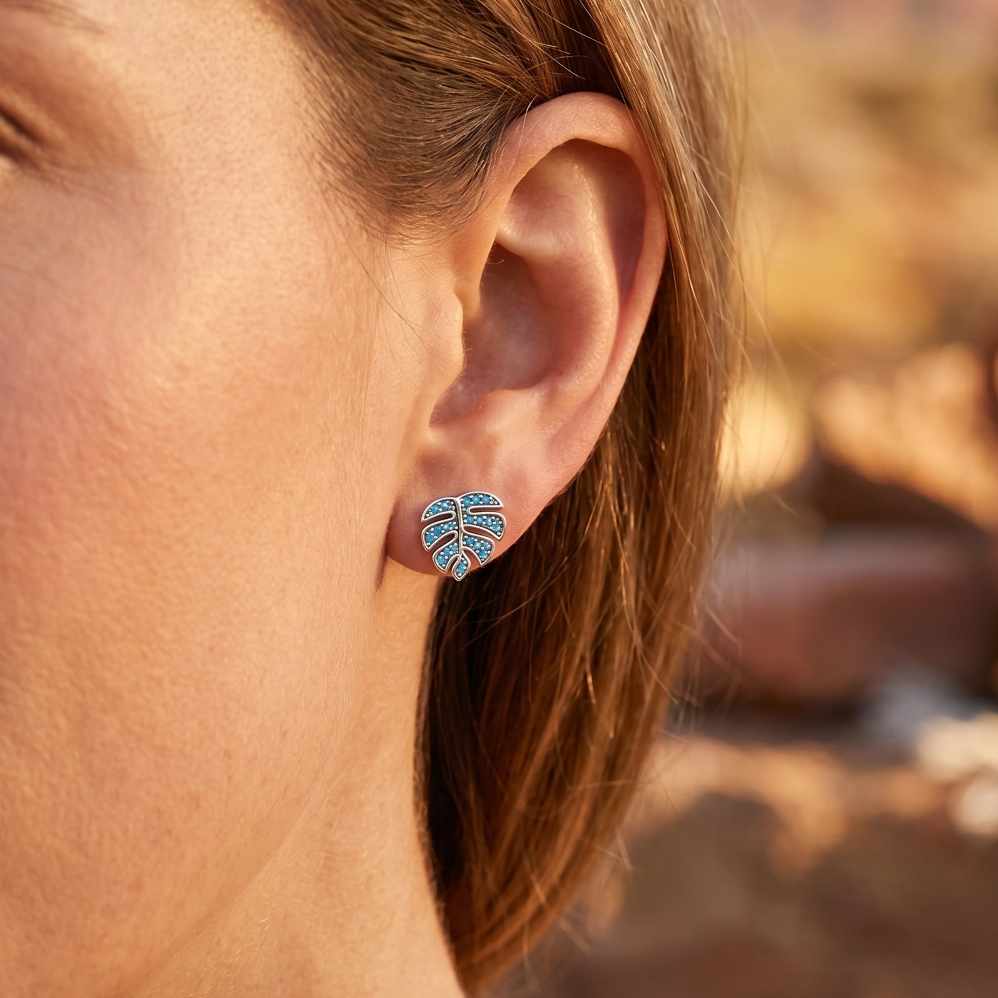 Close-up of a silver monstera leaf earring with turquoise stones worn on a woman's ear.