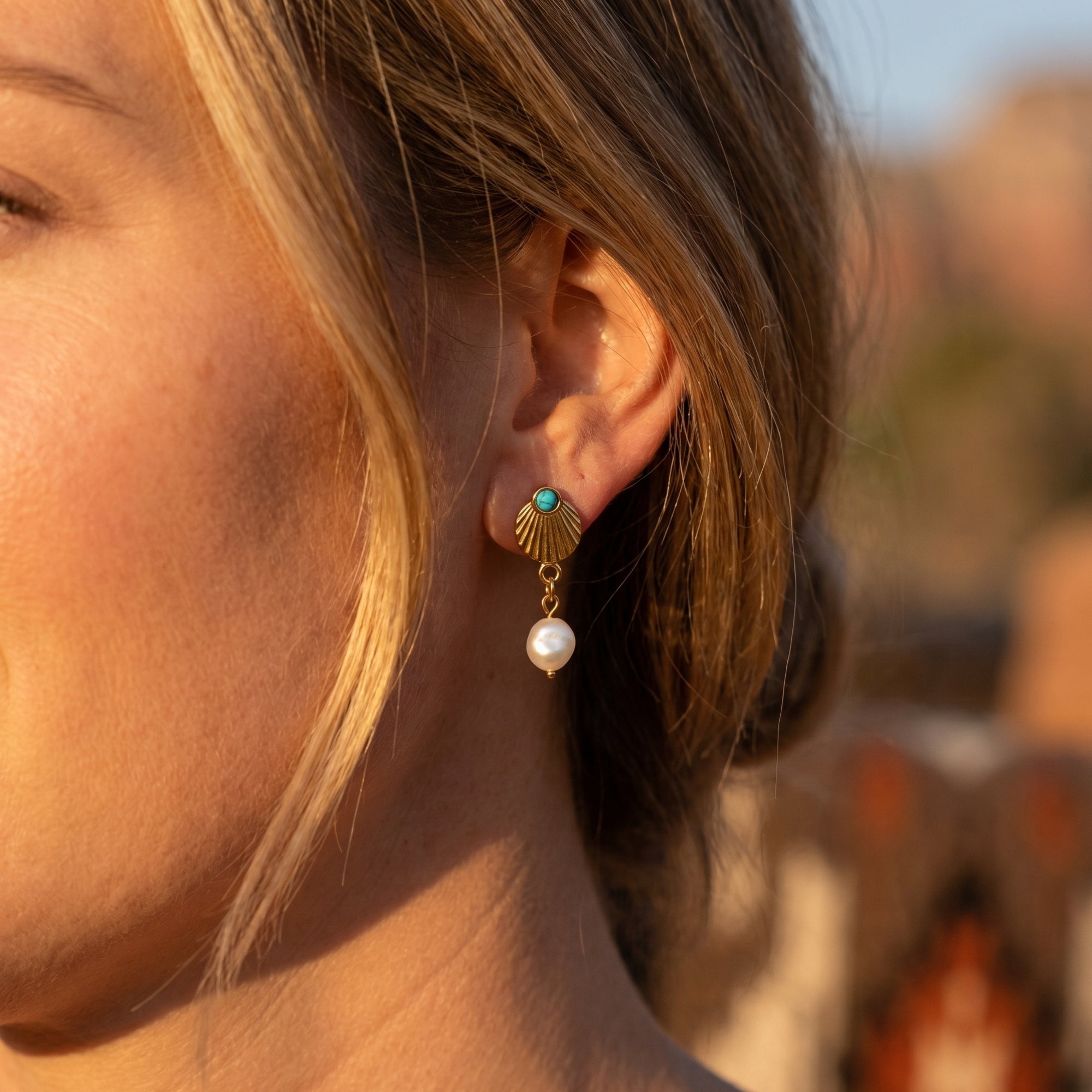 Gold shell drop earring with a turquoise bead and white pearl worn on a model's ear in warm sunlight.