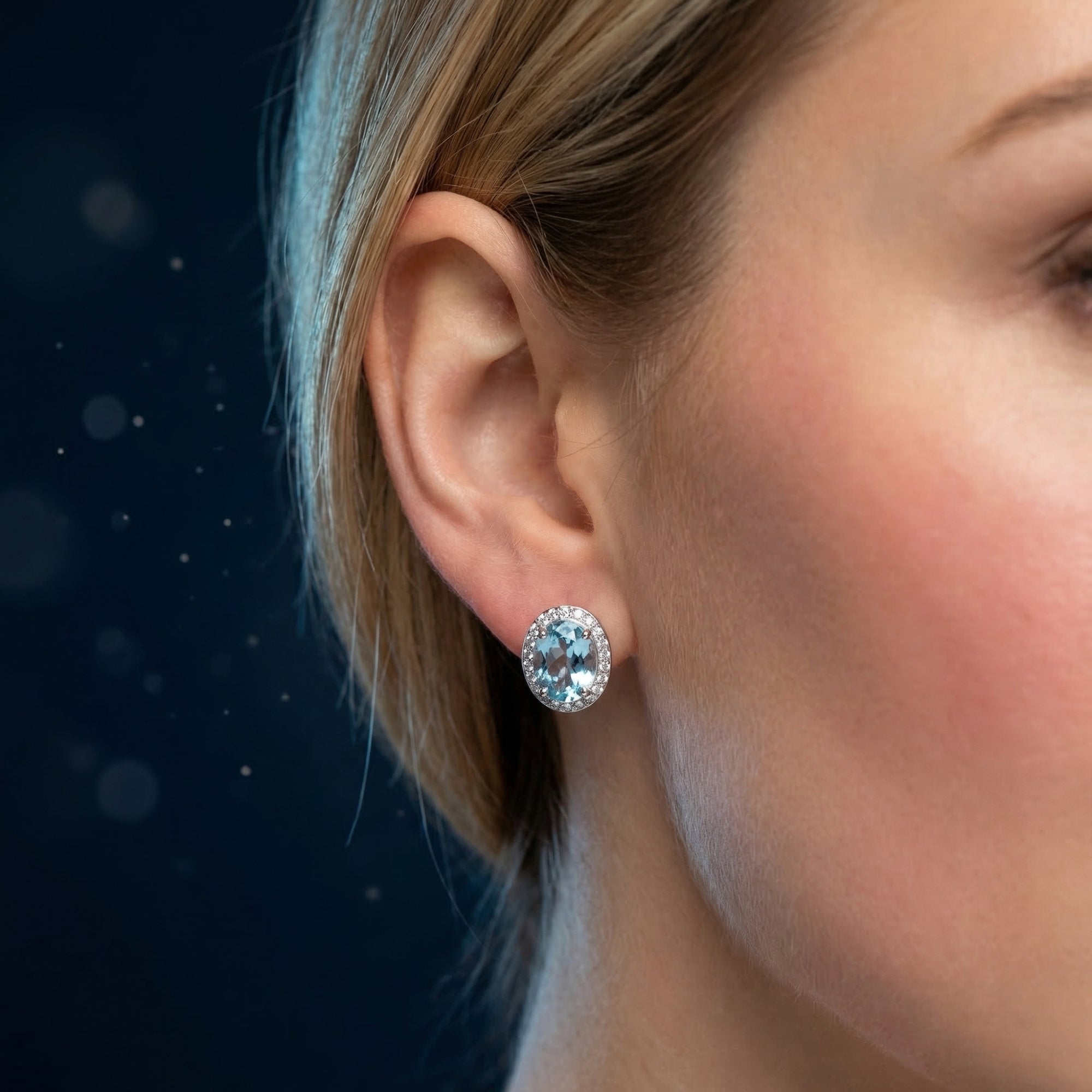 Close-up of a white gold oval blue topaz halo drop earring worn on a model's ear against a dark background.