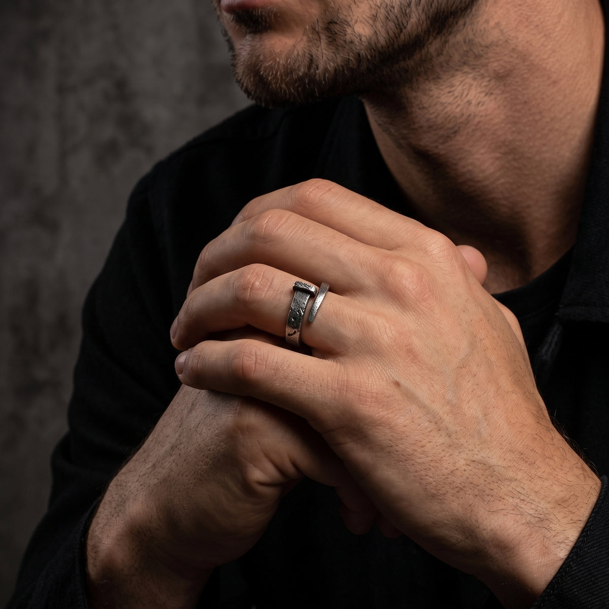 Textured silver nail-style open-band ring worn on a man's finger as his clasped hands rest near his face.