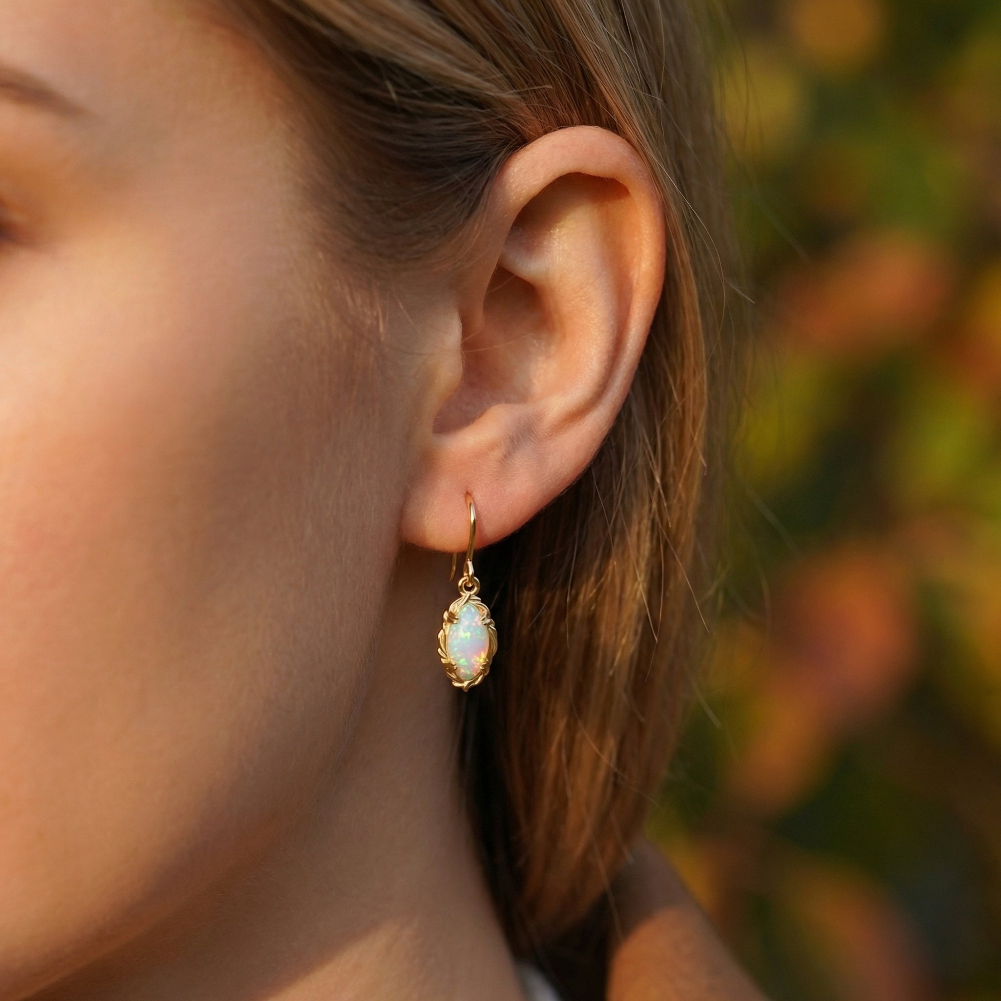 A model wearing a yellow gold drop earring featuring a marquise-cut white opal with iridescent colors.