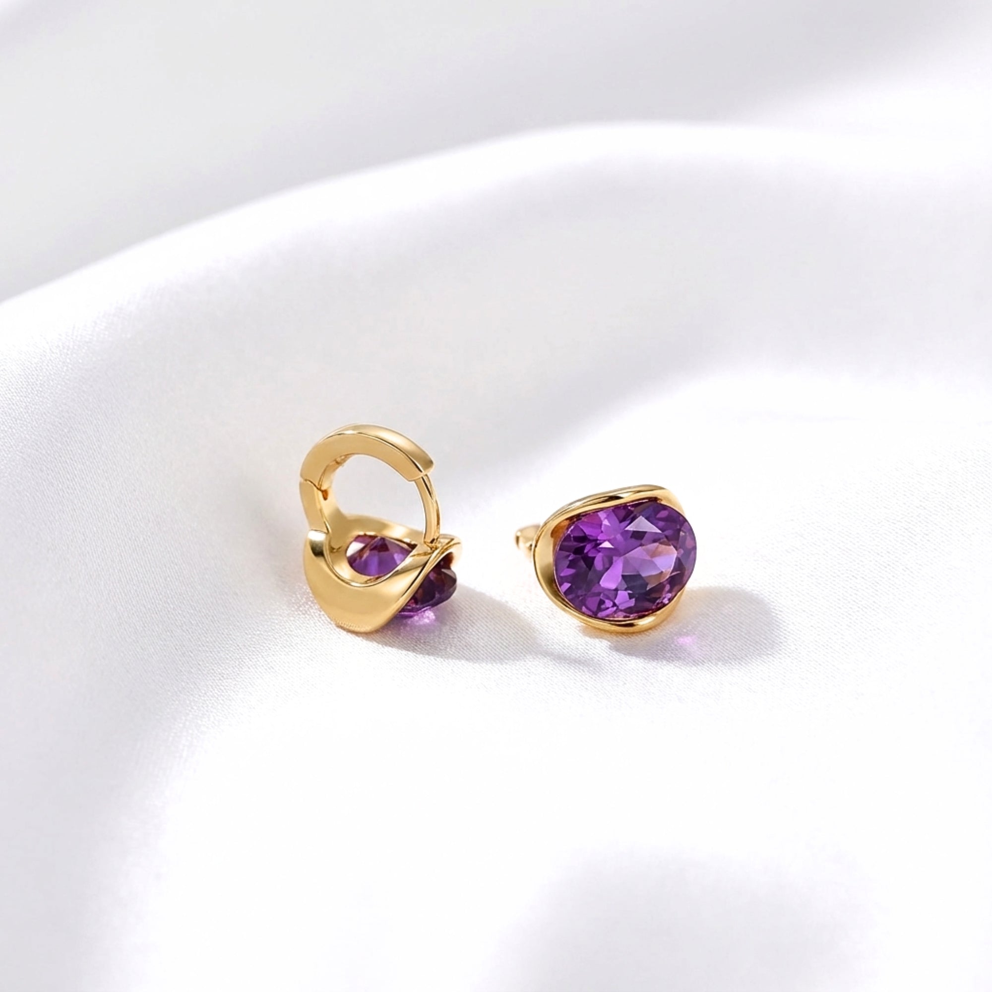 Detail view of 18K gold vermeil hoop earrings with oval cut simulated amethyst stones on white fabric.