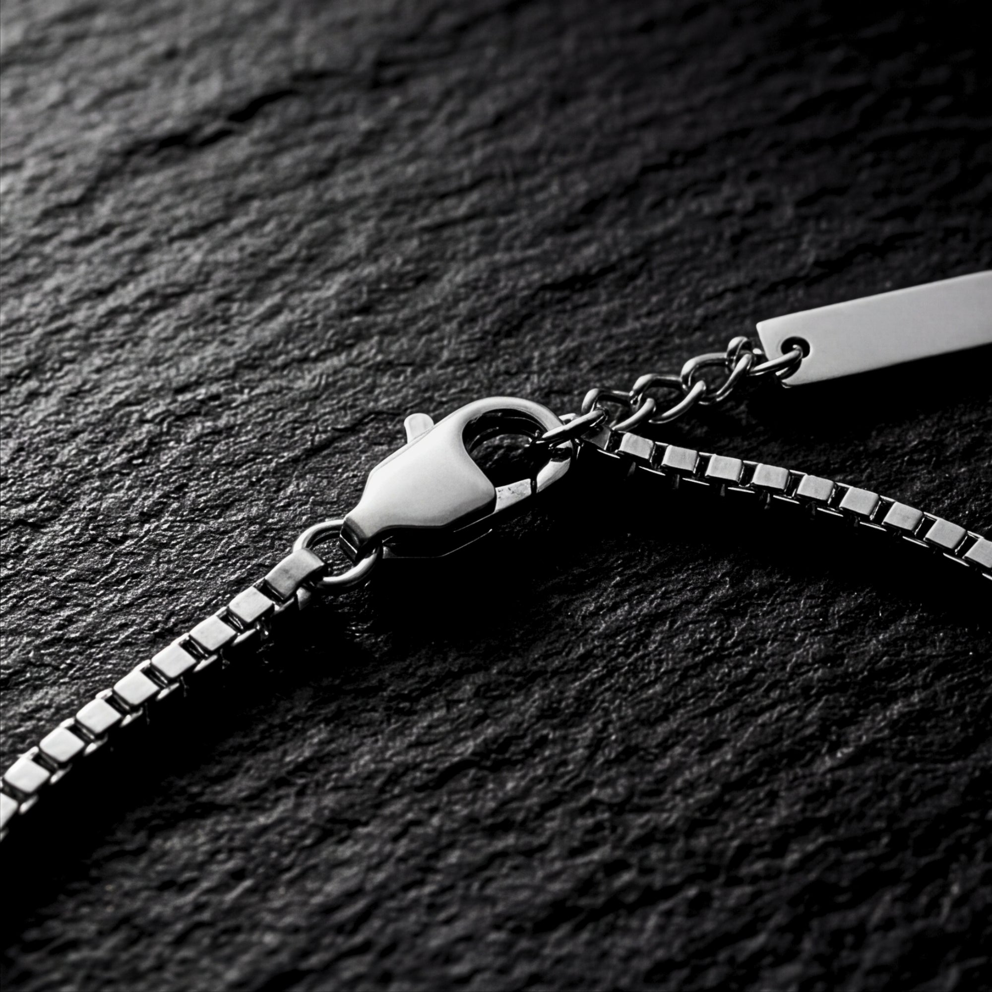 Close-up of the lobster clasp and silver stainless steel box chain links on a dark textured surface.