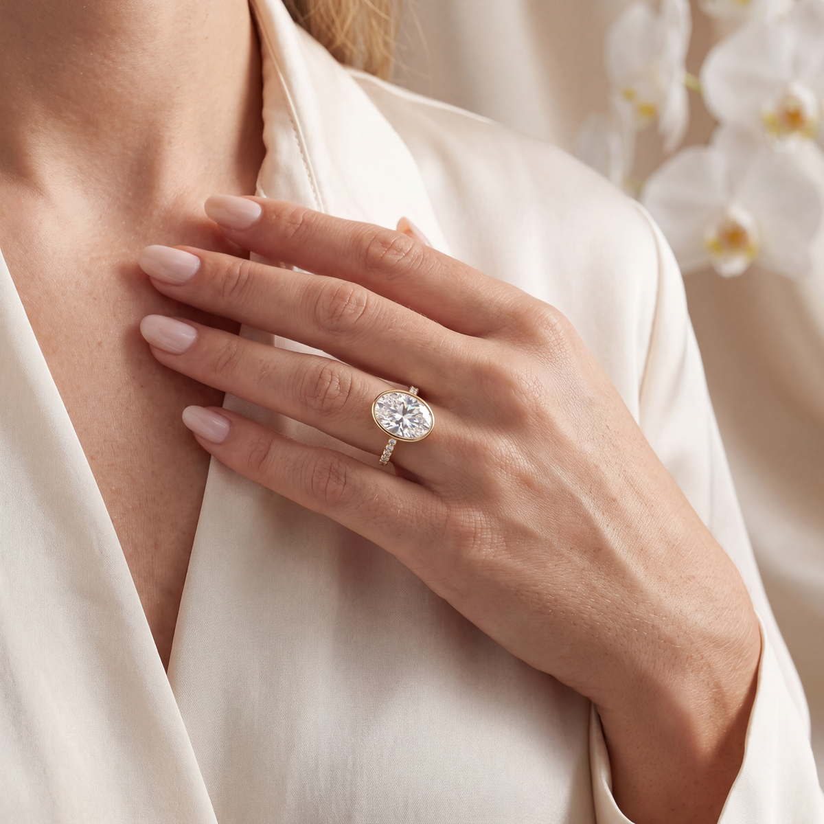 A model wearing an oval moissanite bezel engagement ring in yellow gold resting her hand on her chest.