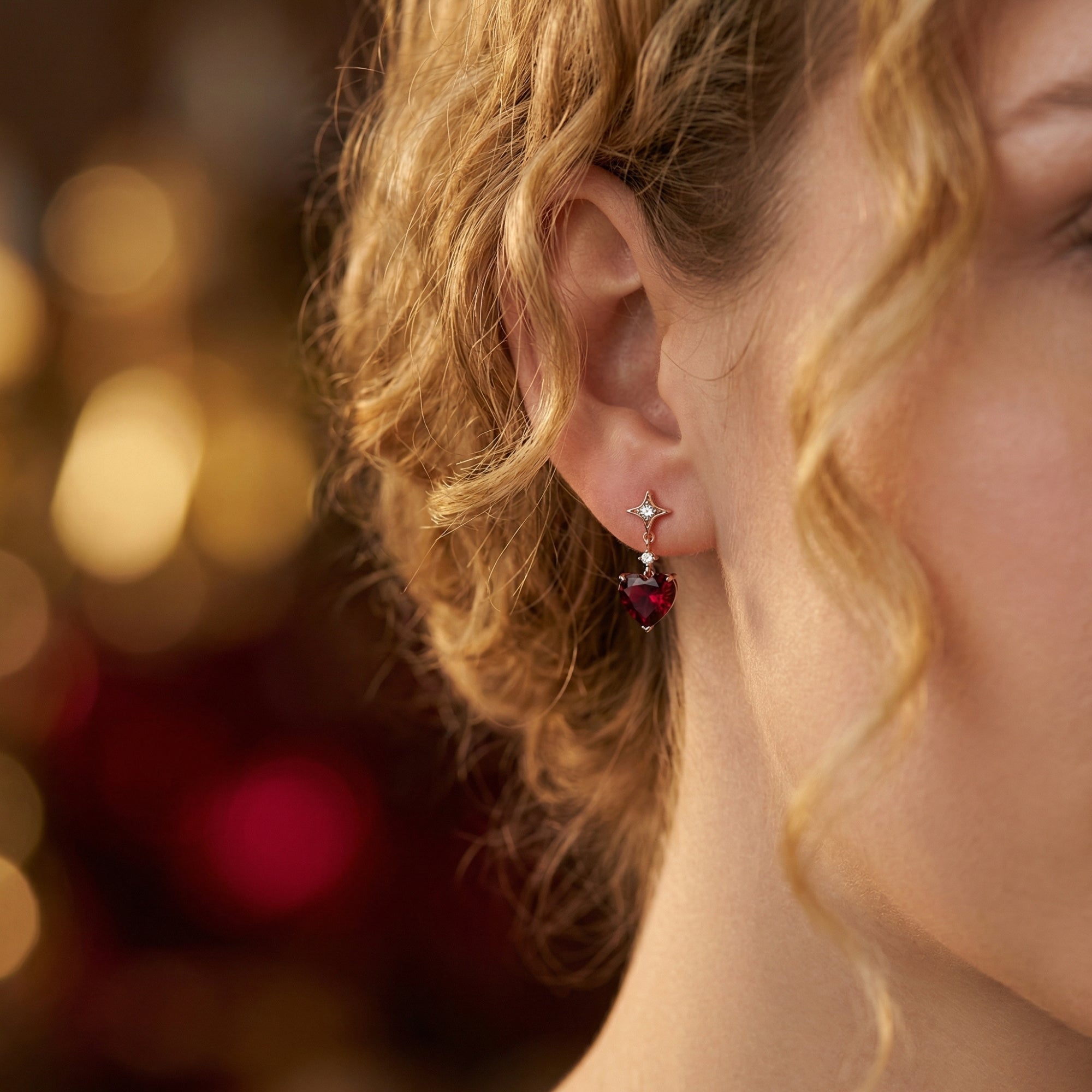 Side view of a woman wearing a rose gold heart cut garnet drop earring against a warm bokeh background.