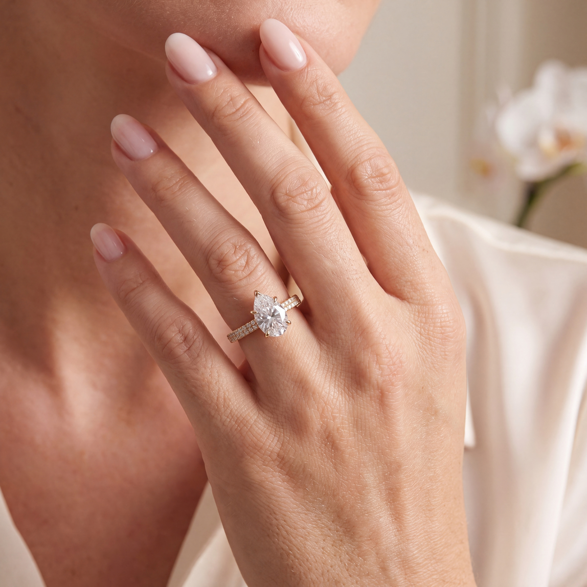Pear-cut moissanite ring with a double-row pavé gold band worn on a hand near the face.