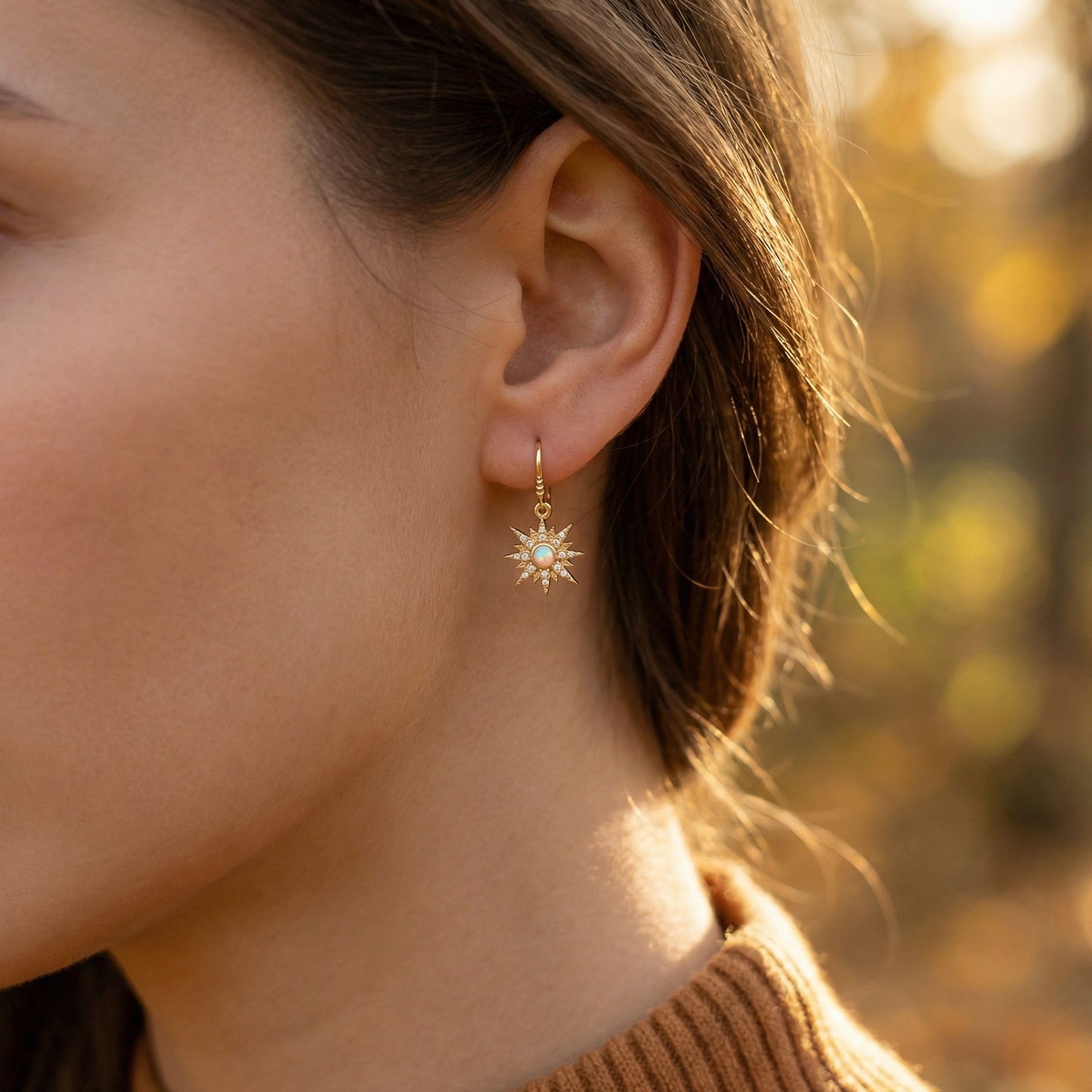 Gold-tone starburst drop earring with a round white opal worn on a model's ear in warm outdoor light.