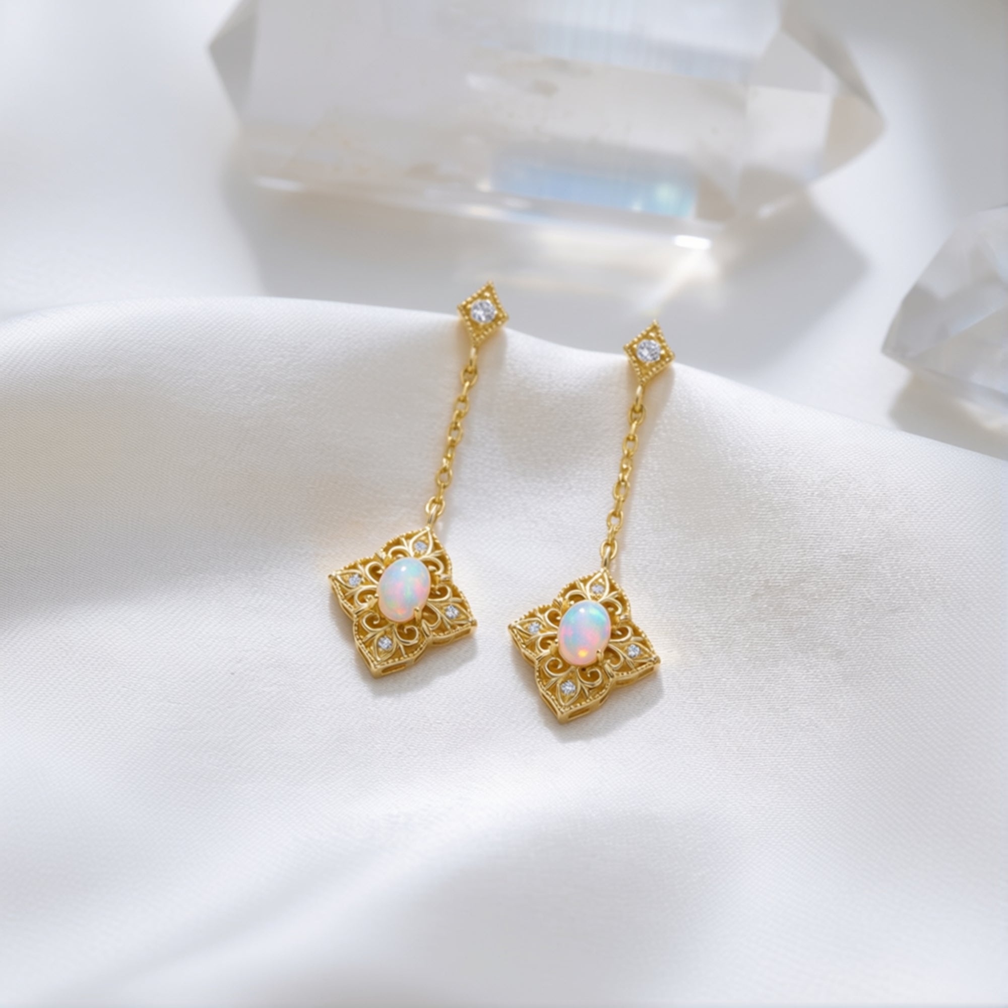 Two gold-tone filigree drop earrings with oval white opal stones resting on white silk fabric.