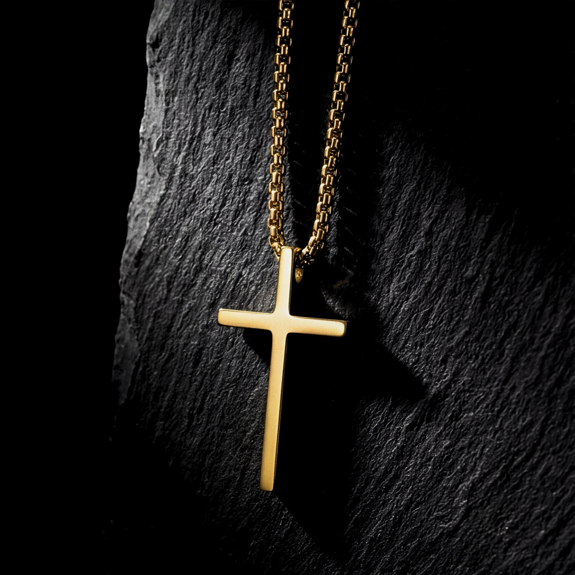 Gold tone stainless steel Devotion Cross necklace hanging vertically against a dark, rugged stone background.