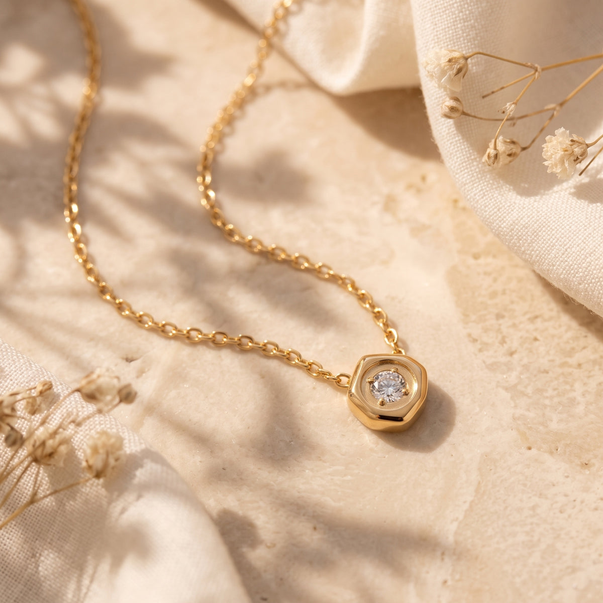 Angled close-up of the gold-tone hexagon pendant and clear stone resting on a beige stone surface.