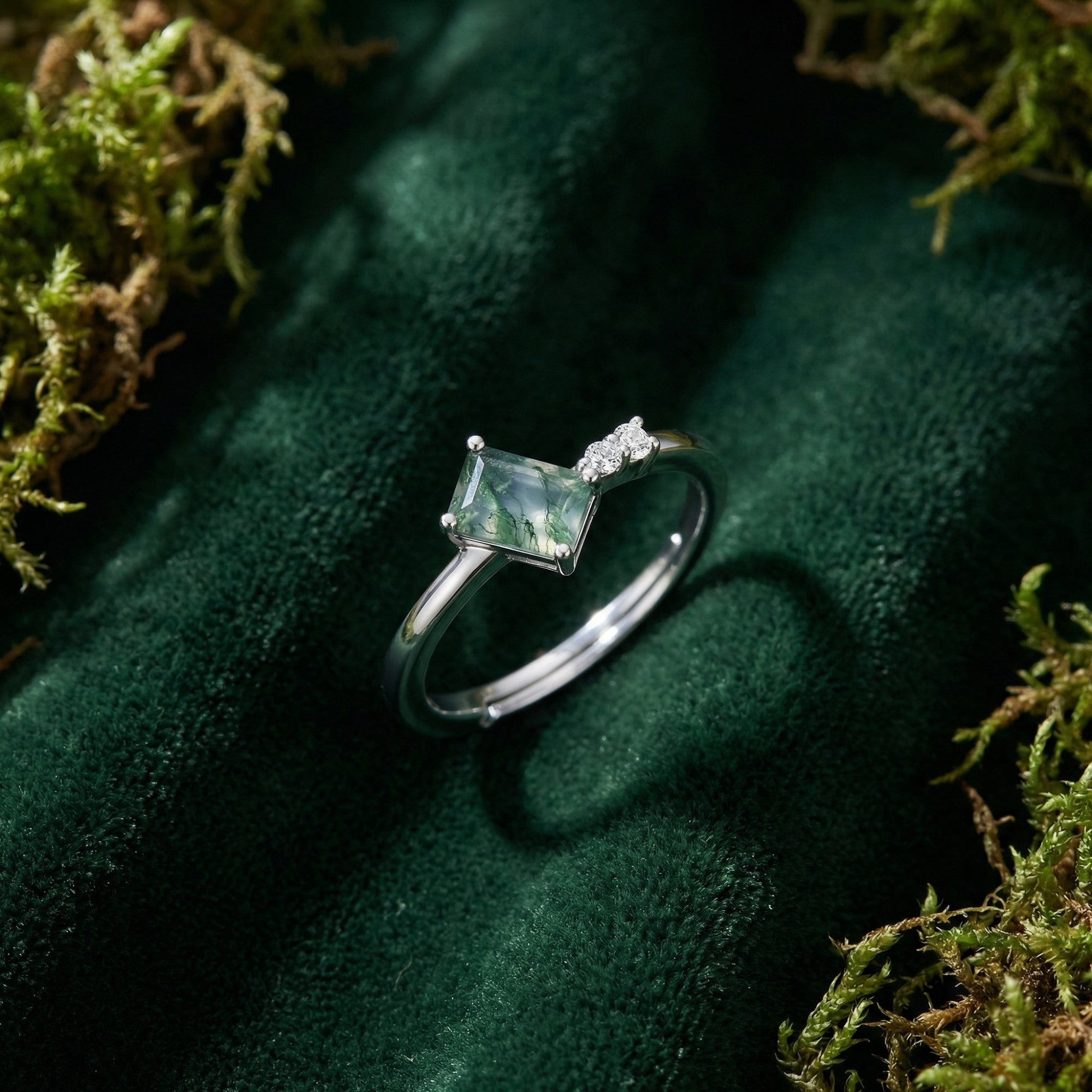 Close-up of the adjustable rectangular moss agate ring with side accents on a green mossy fabric background.