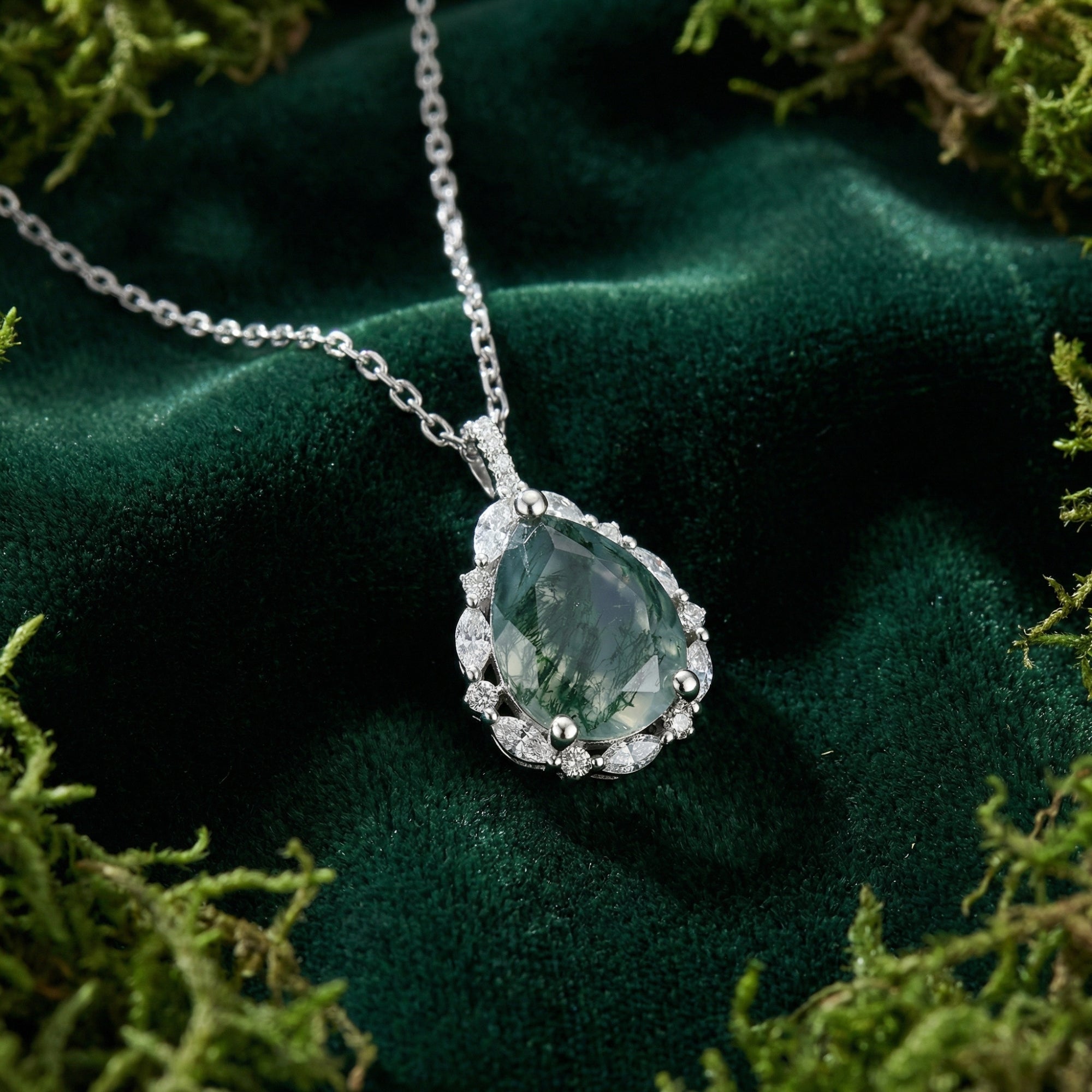 Close-up of a pear-cut Moss Agate pendant with sterling silver halo setting on a delicate chain over green velvet.