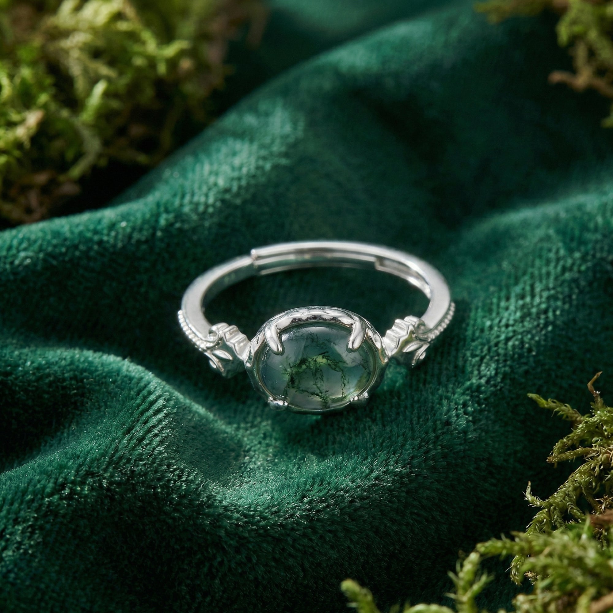 Close-up of an oval natural moss agate ring in S925 sterling silver with leaf details on green velvet.