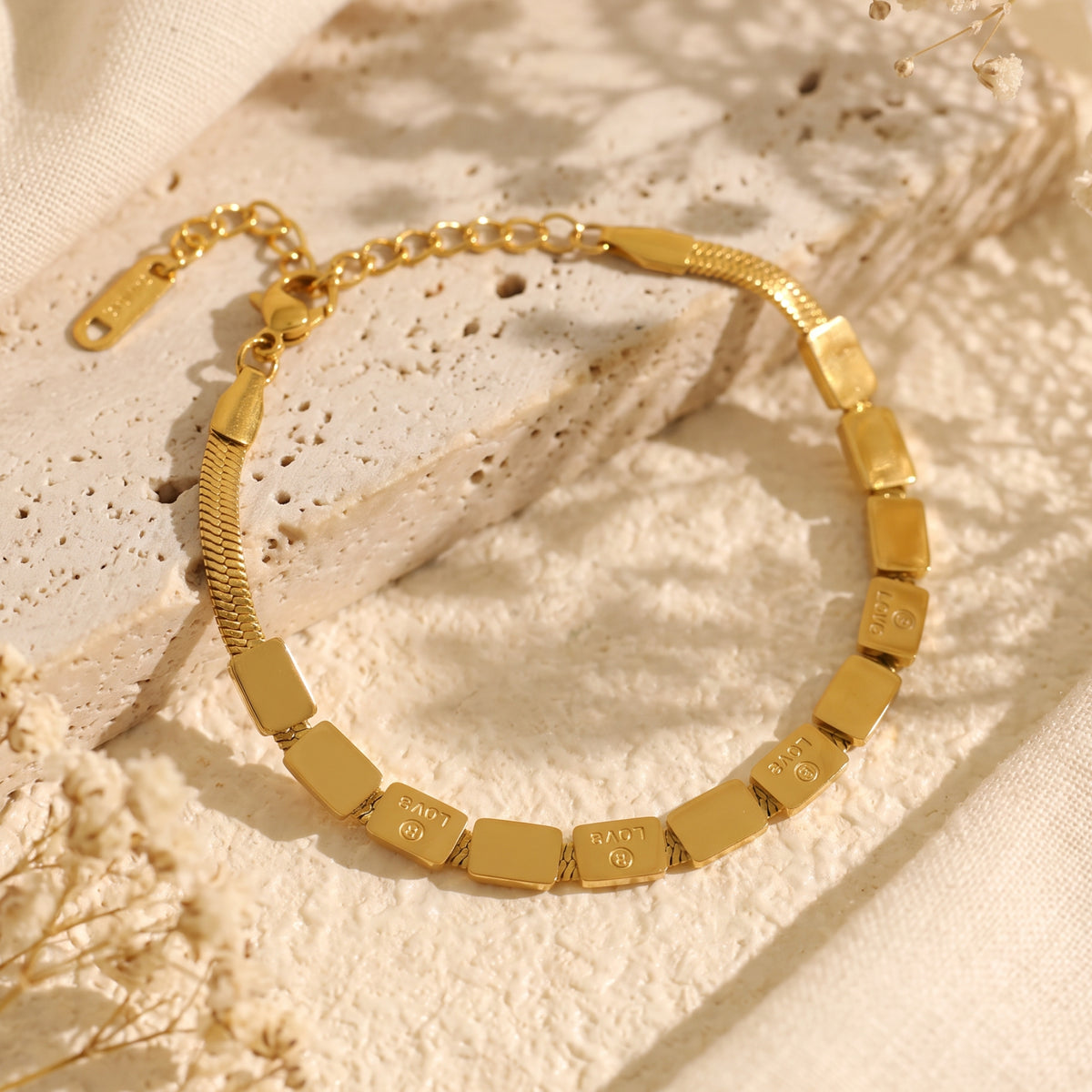 Gold-tone inscribed tile bracelet laid out in a loop on a beige stone surface.