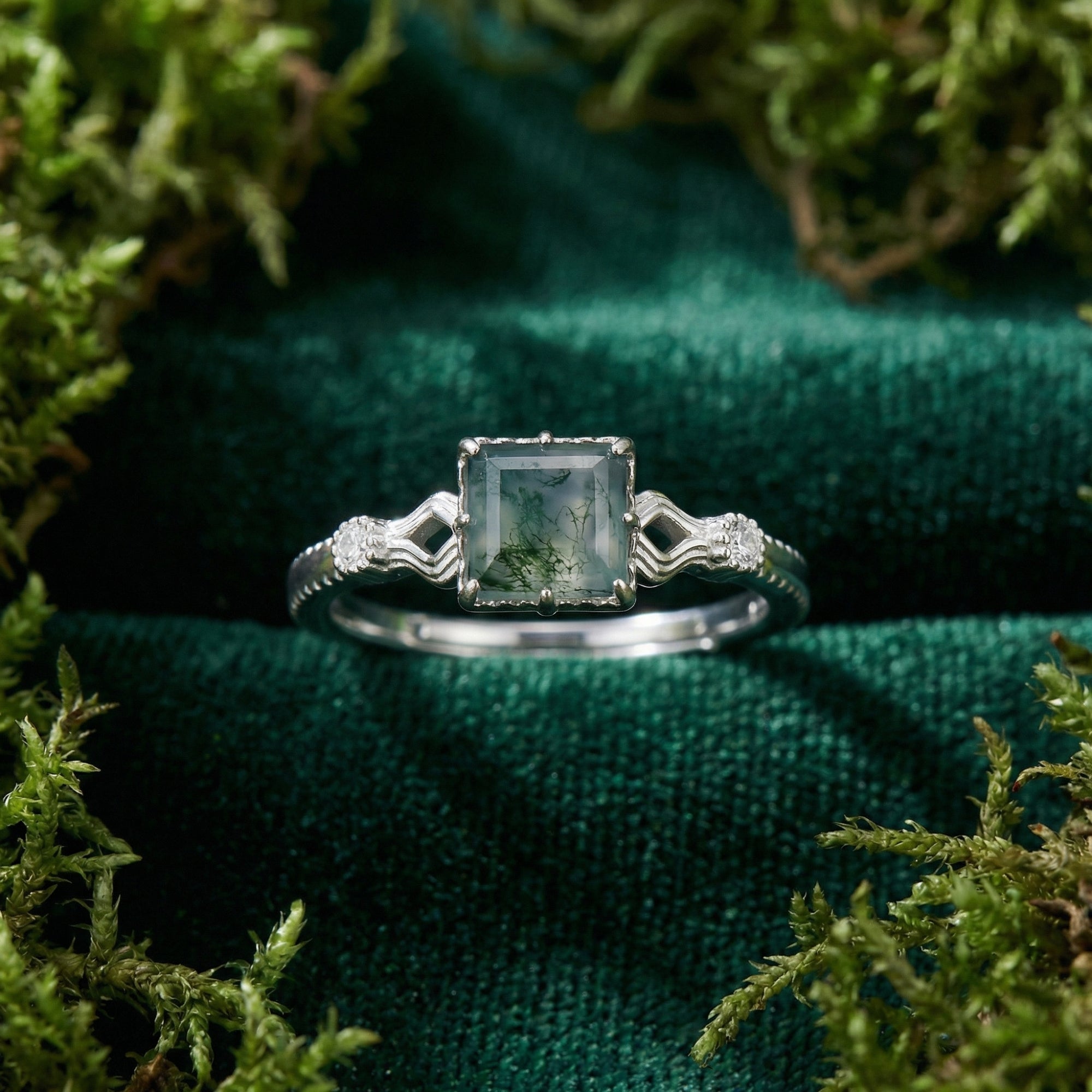 Close-up of a cushion cut moss agate stone set in a sterling silver wavy band ring on a green velvet surface.