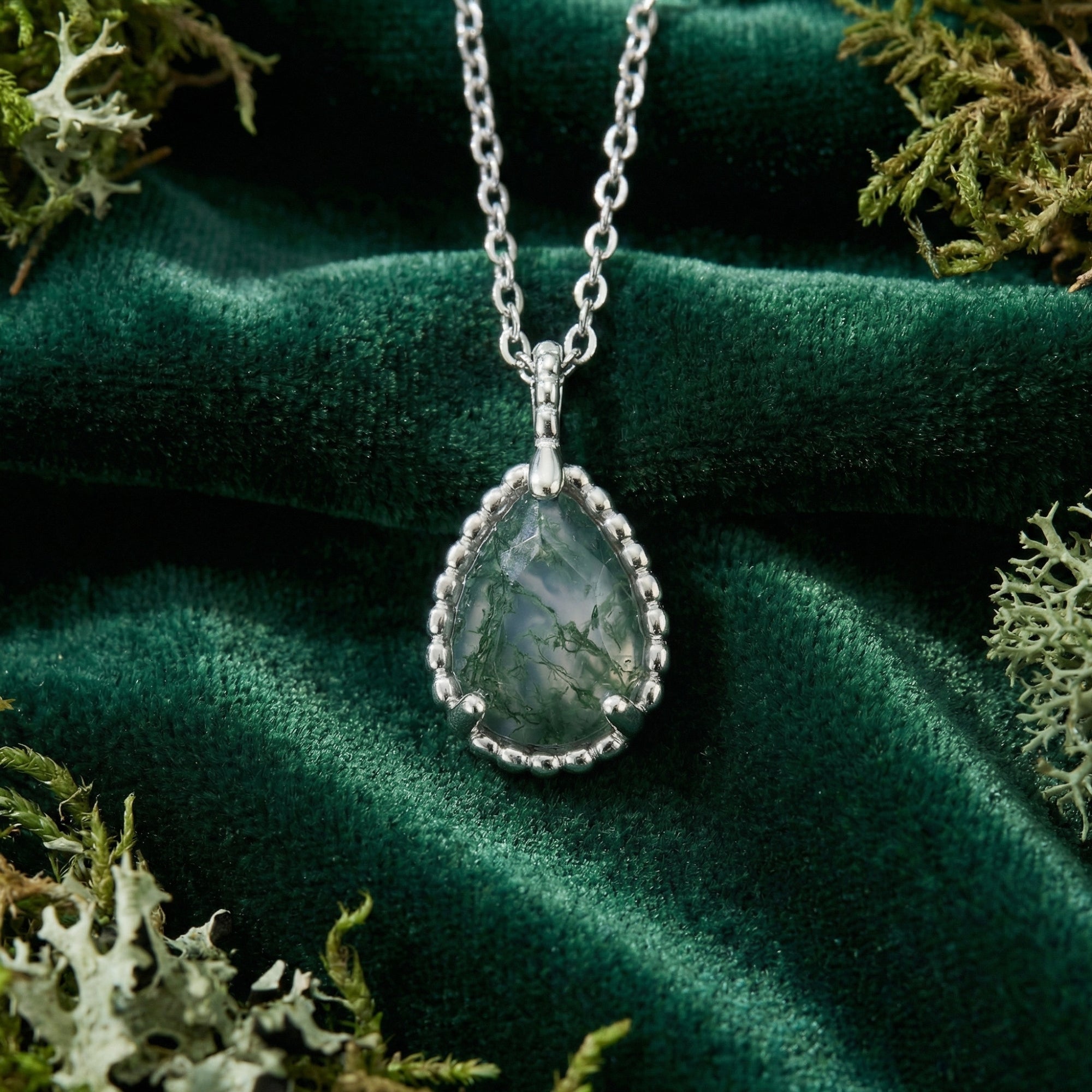 Macro view of the teardrop moss agate stone and silver basket weave setting on a green velvet background.