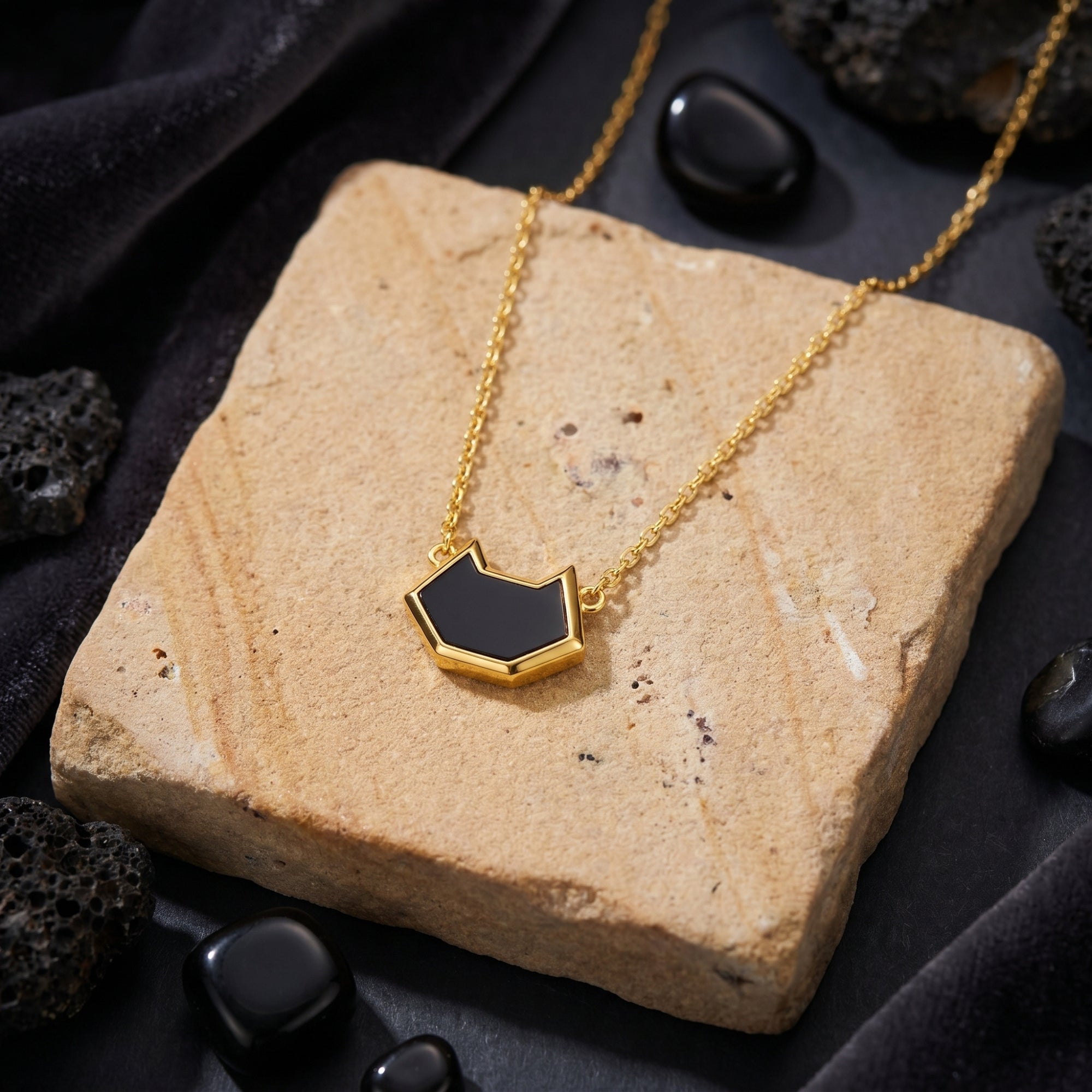 Close-up of the irregular-cut black onyx pendant and 18K gold plated chain resting on a textured stone surface.